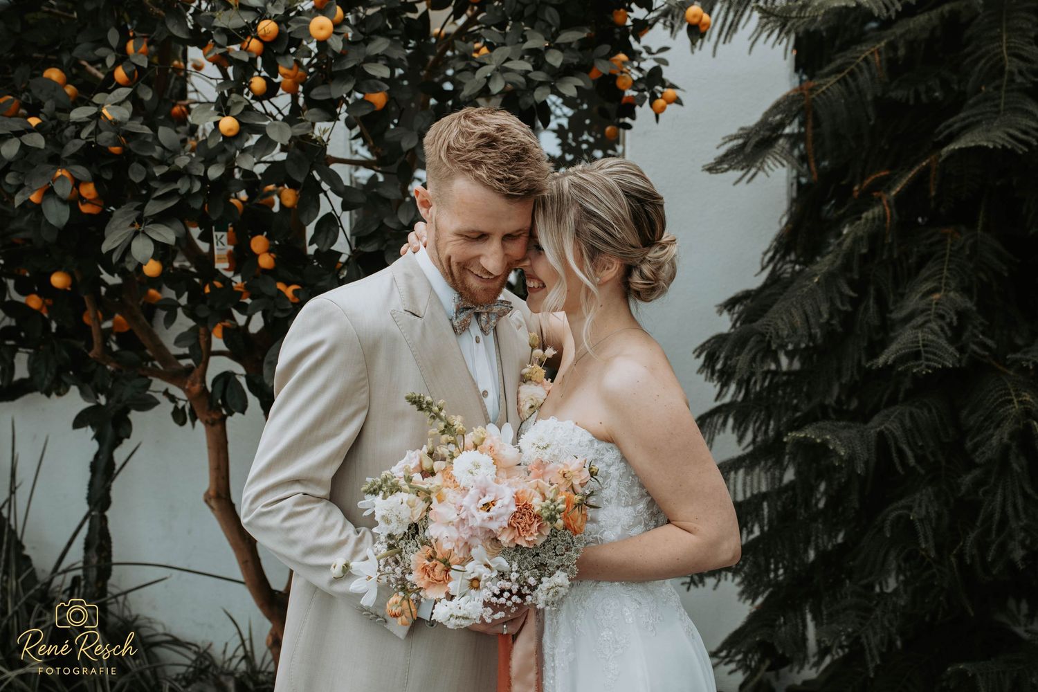 Bride and groom share intimate moment under orange tree, bride holding pastel bouquet in lush garden wedding setting.