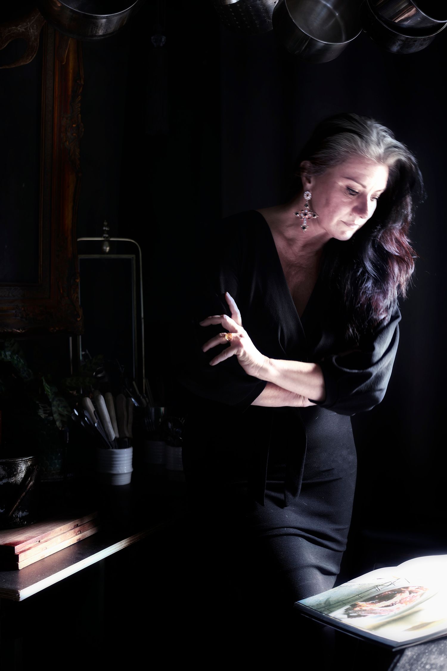 Woman in a black dress with her long dark hair down. Arms folded as she leans over her black kitchen table to read the recipe book