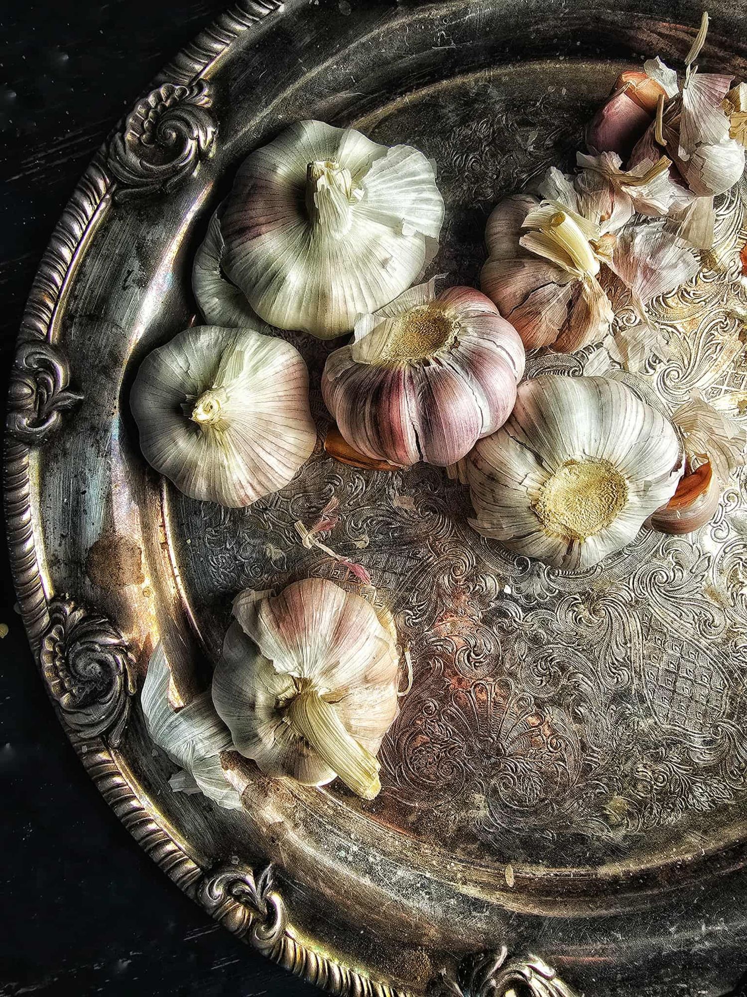 A photo of an ornate vintage silver tray scattered with garlic bulbs and garlic cloves.