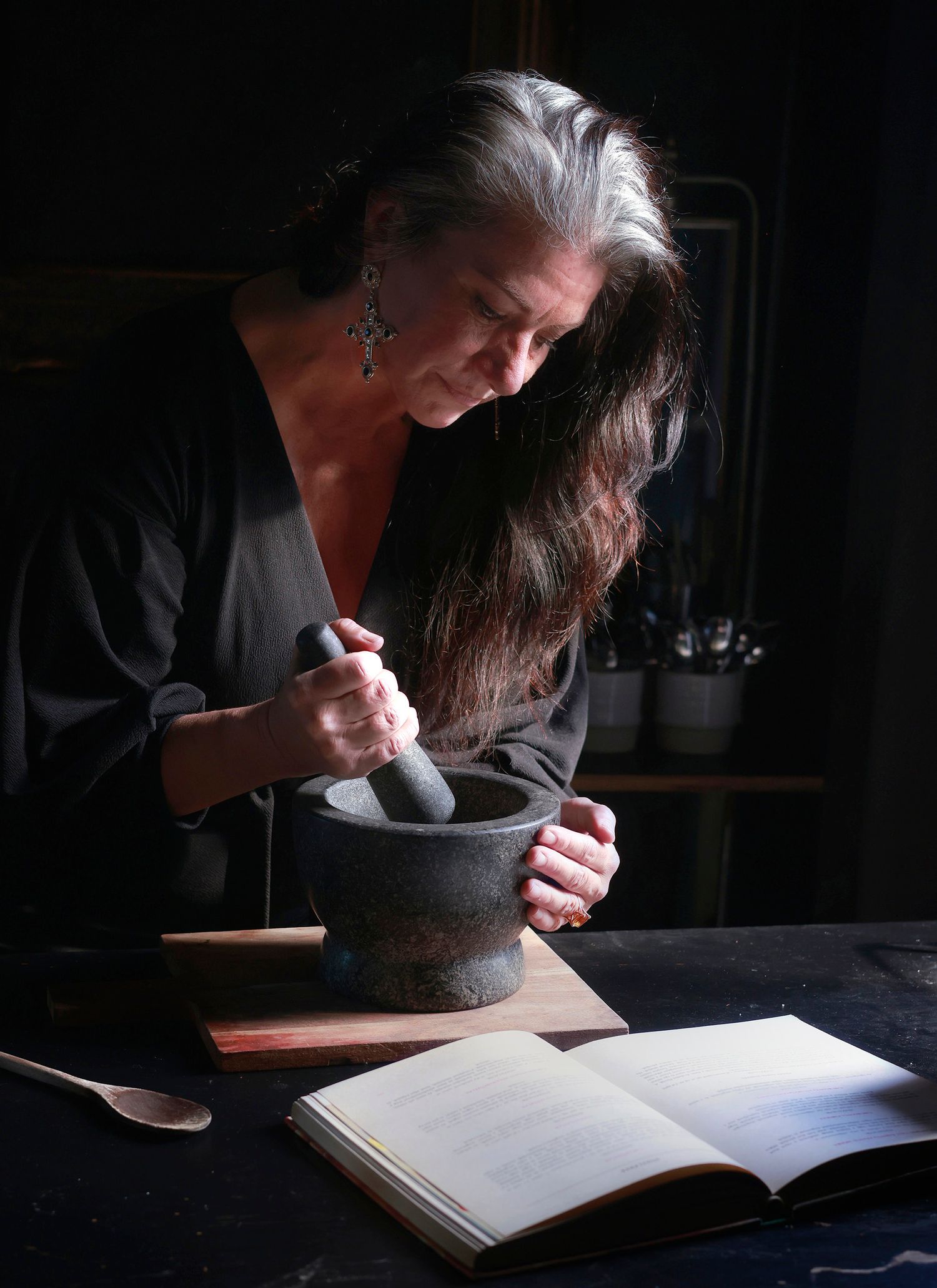 Photo of Ilona working a pestle and mortar while looking at an open cookbook in front of her.