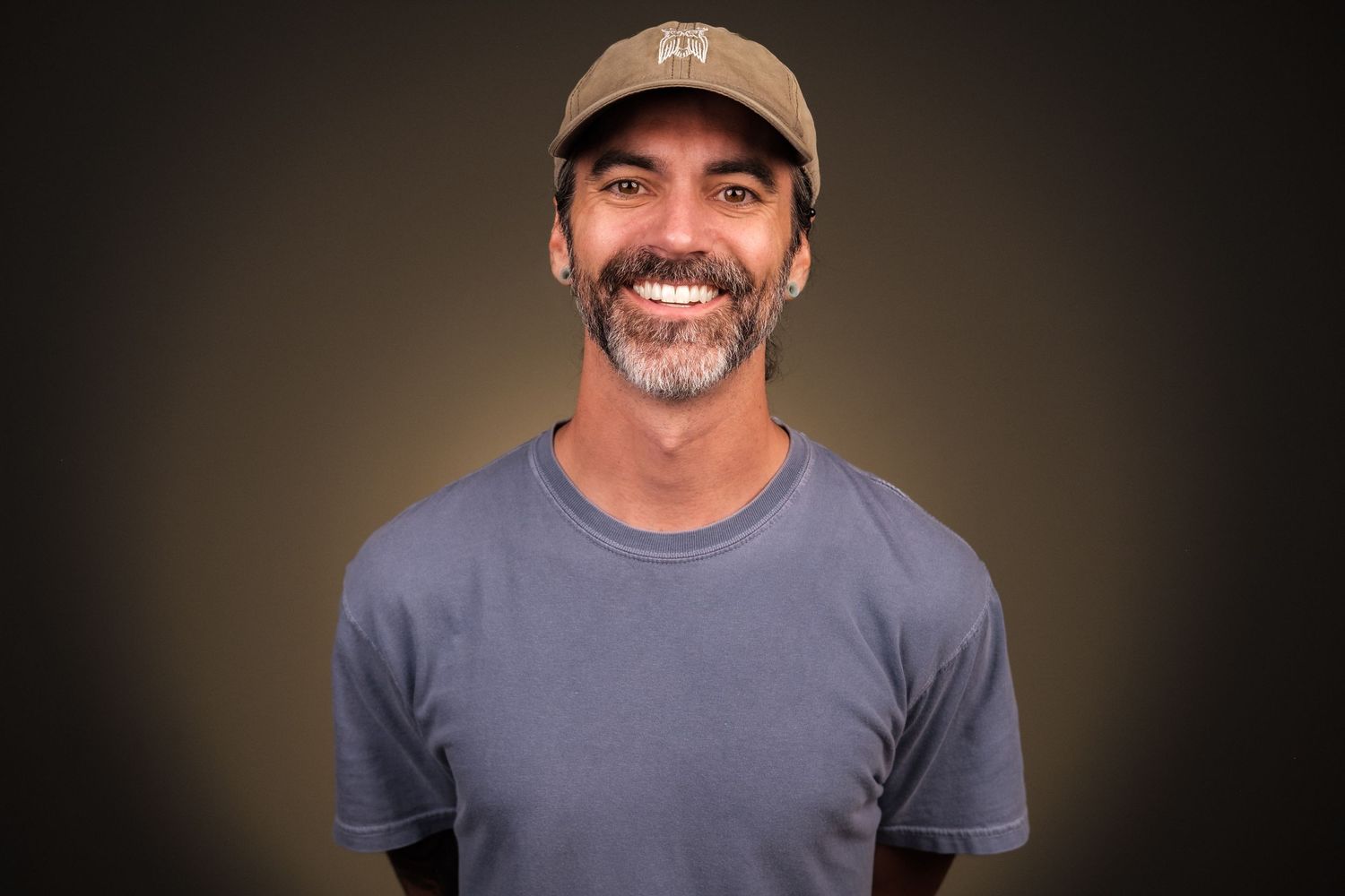 Casual professional headshot of smiling man wearing hat and blue shirt against a warm studio background, photographed in Huntsville, Alabama