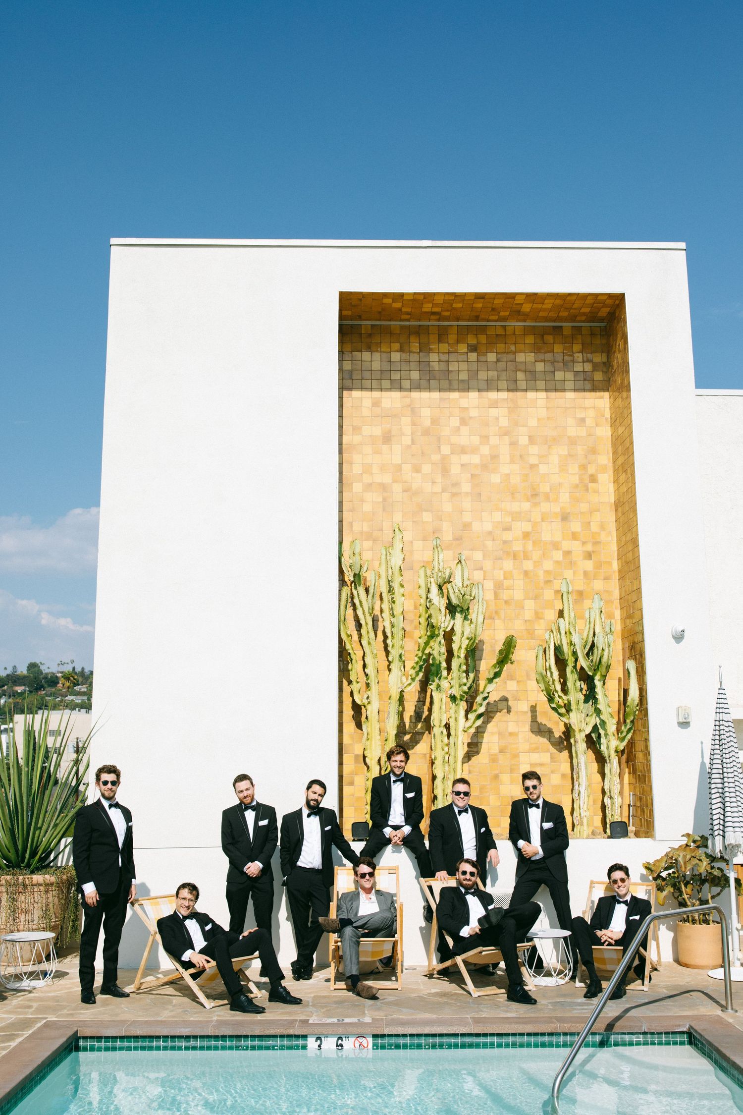 Wedding party in black tuxedos pose creatively against modern beige wall by pool in desert setting.
