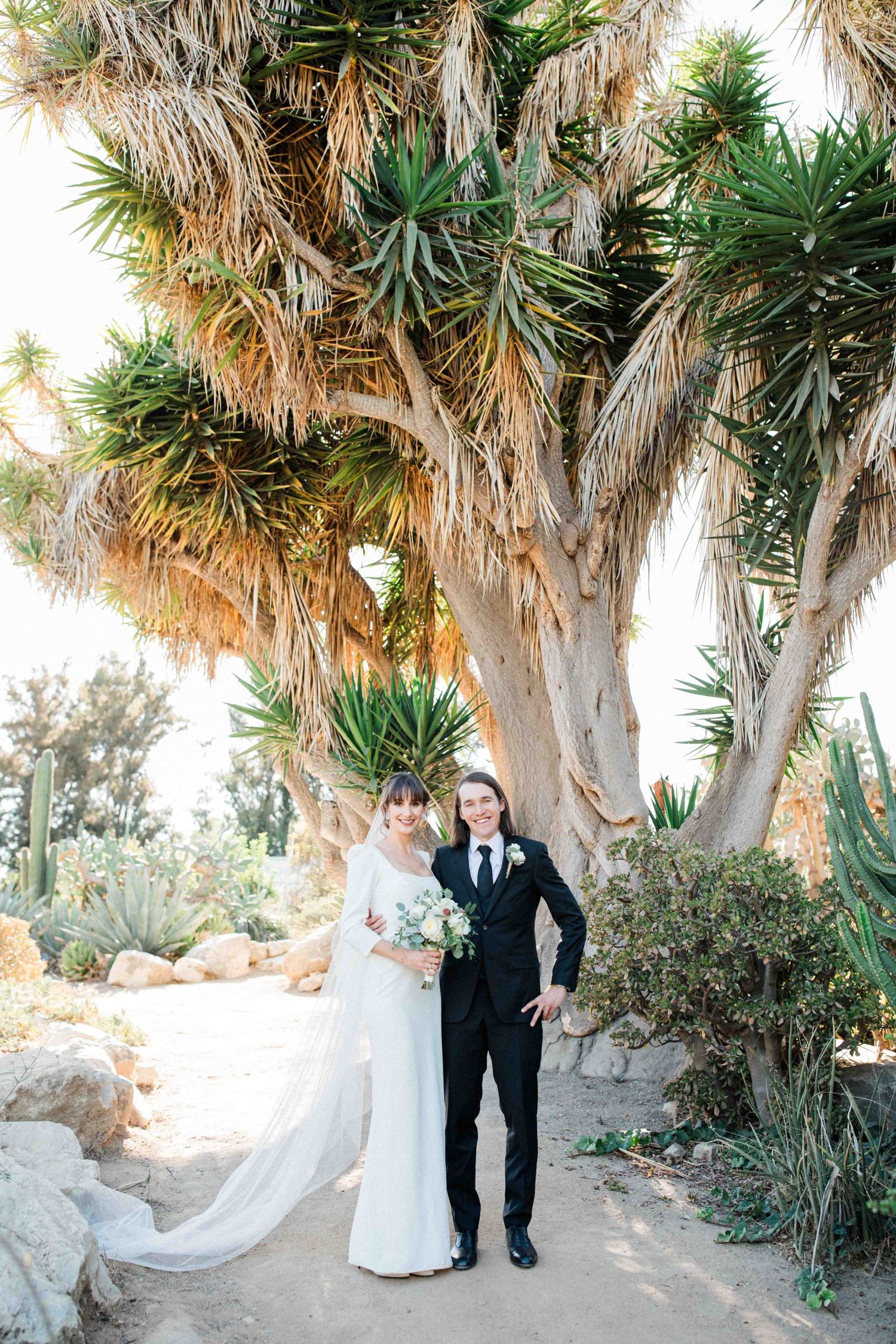 Bride and groom standing together outdoors beneath tall trees on their wedding day