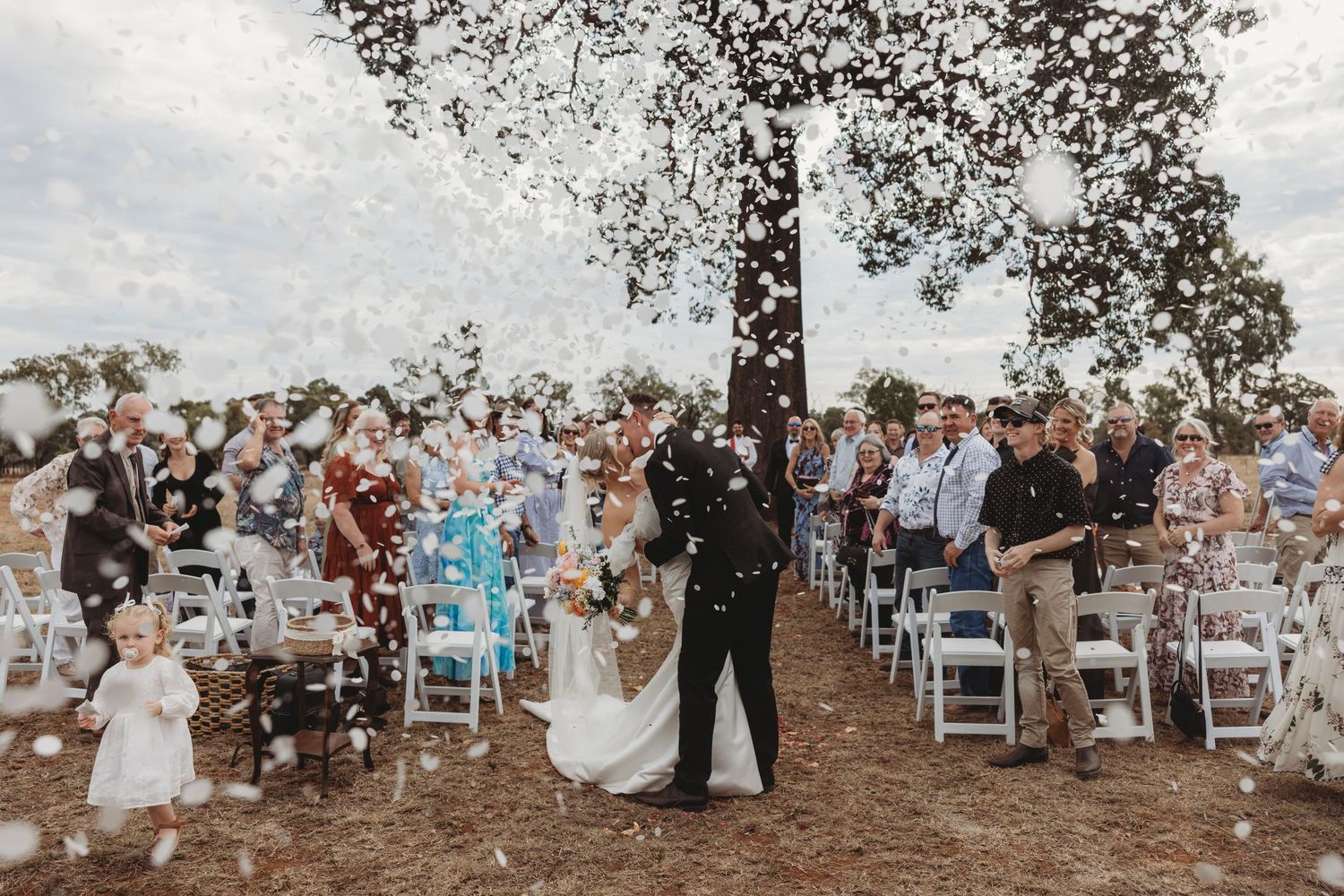 Wedding guests celebrate and cheer during outdoor ceremony with white chairs on rustic grounds.