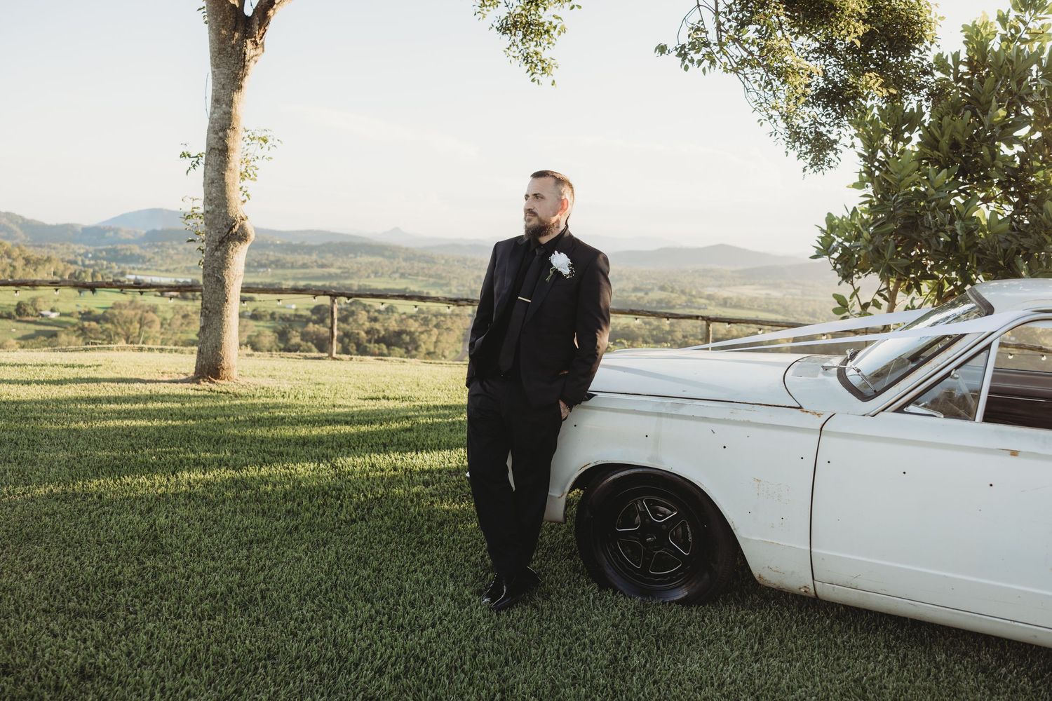 Groom in black suit leaning against vintage white car with countryside backdrop.