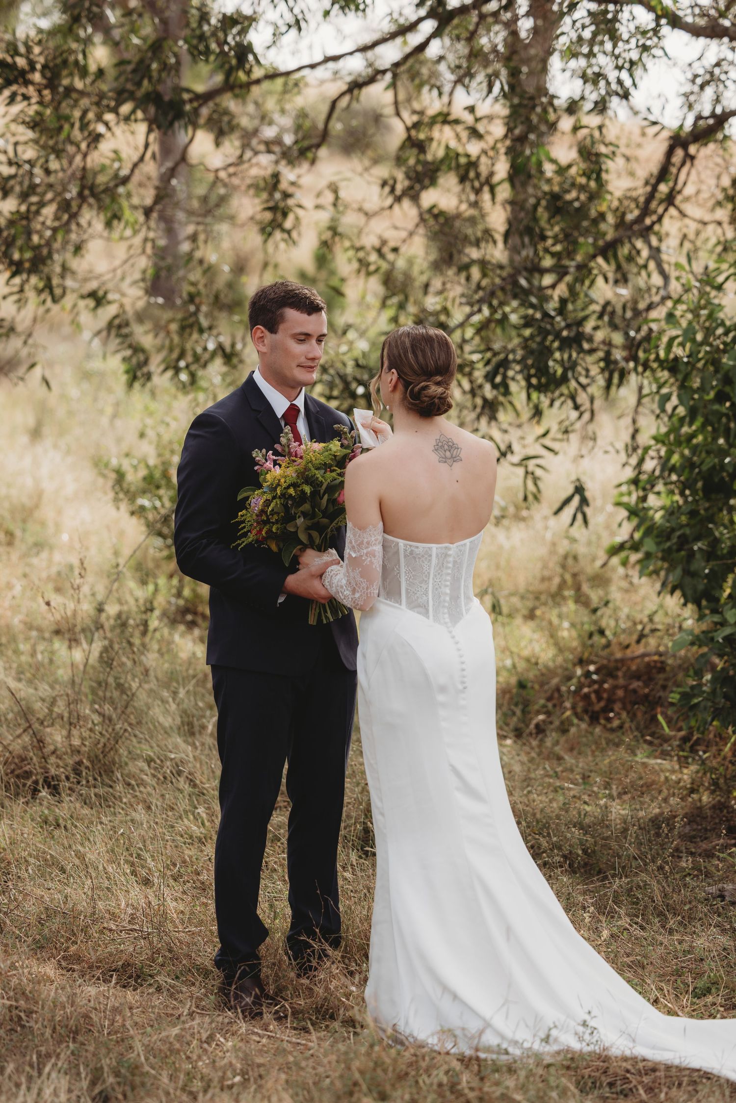 A groom in a navy suit holds a bouquet facing his bride in a white lace wedding dress outdoors.