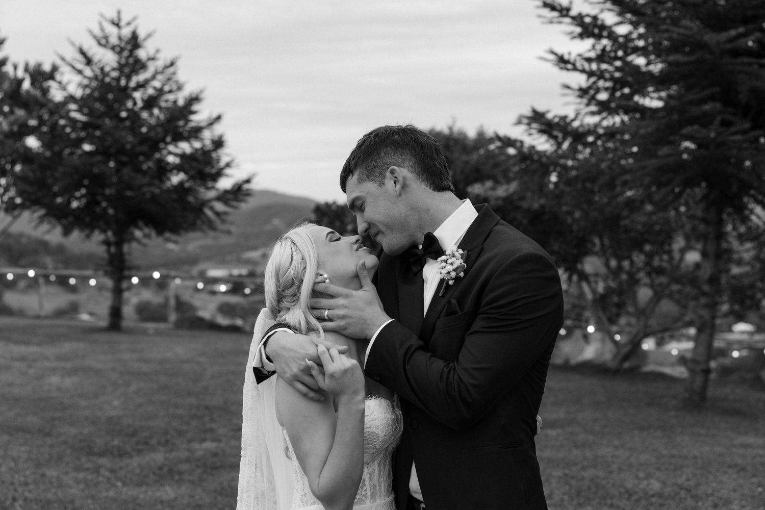 Black and white photo of a bride and groom sharing a kiss outdoors with trees and string lights in the background.