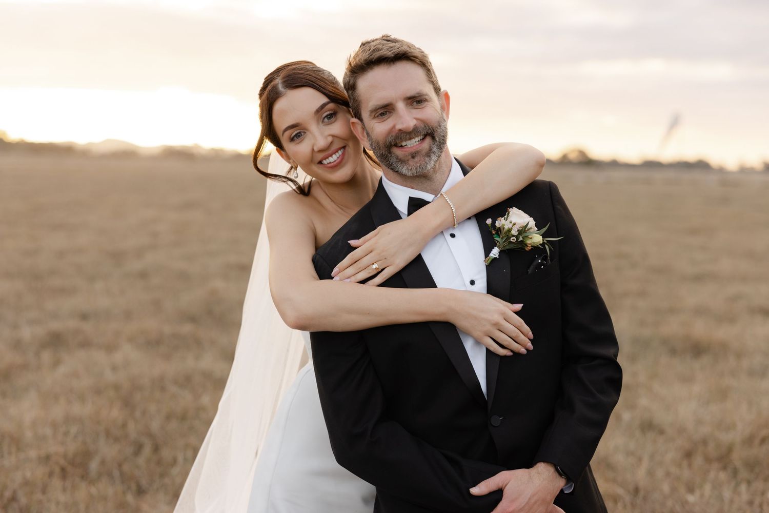A smiling bride and groom embrace in a golden field at sunset, wearing wedding attire with a boutonniere.