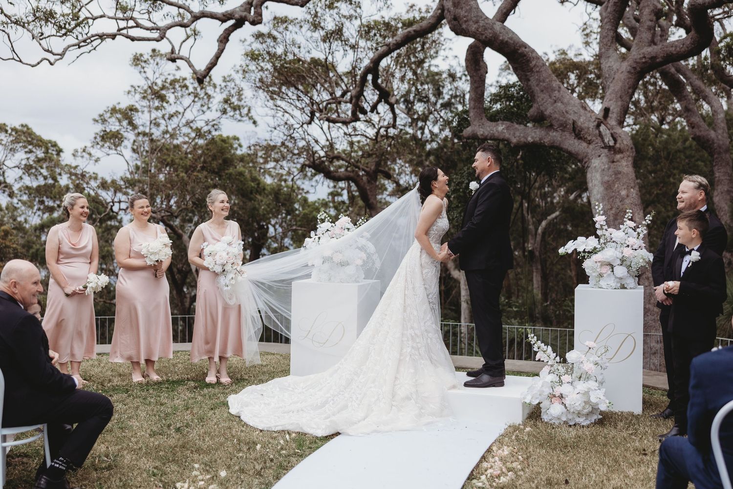 Outdoor wedding ceremony under a large tree with bridesmaids in pink dresses and a flowing white veil in the breeze.