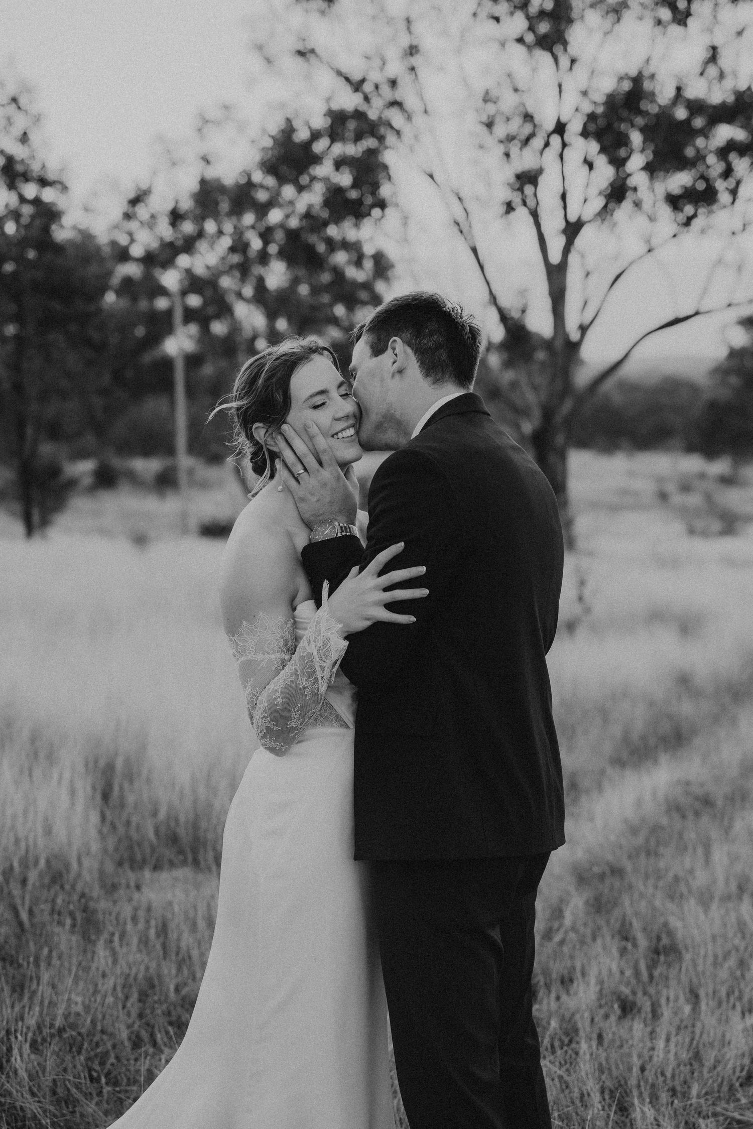 Black and white photo of groom kissing bride on the cheek in a field at dusk during wedding.