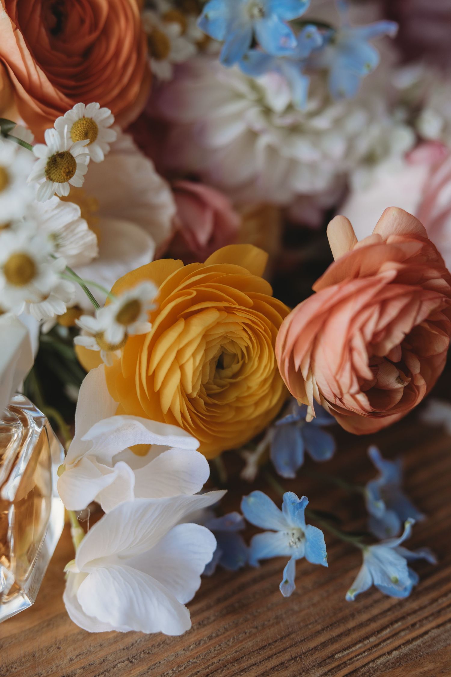Romantic floral arrangement with orange ranunculus, pink roses and white daisies on wooden surface.