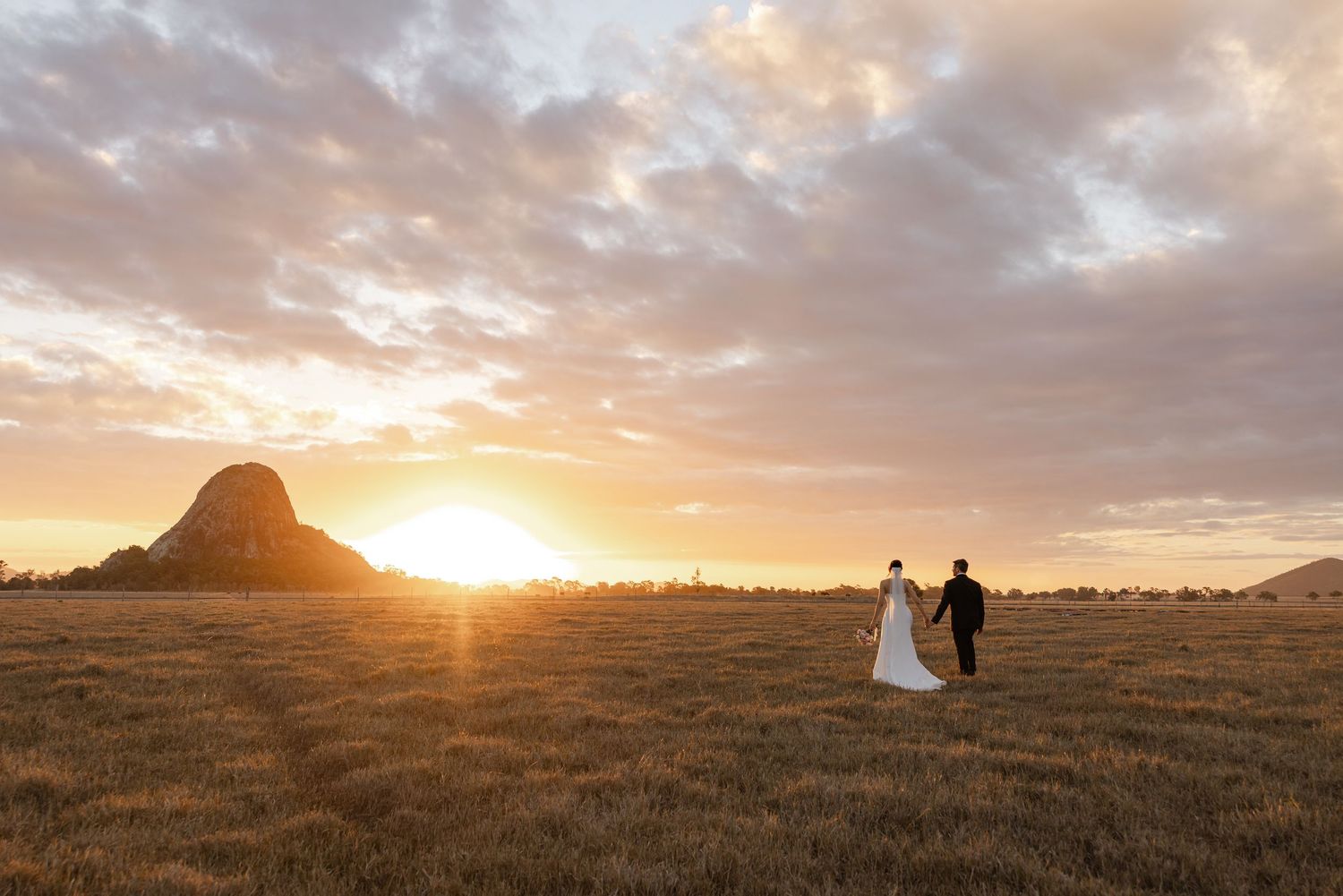 Bride and groom walk hand in hand through golden fields at sunset with a dramatic rocky mountain backdrop.