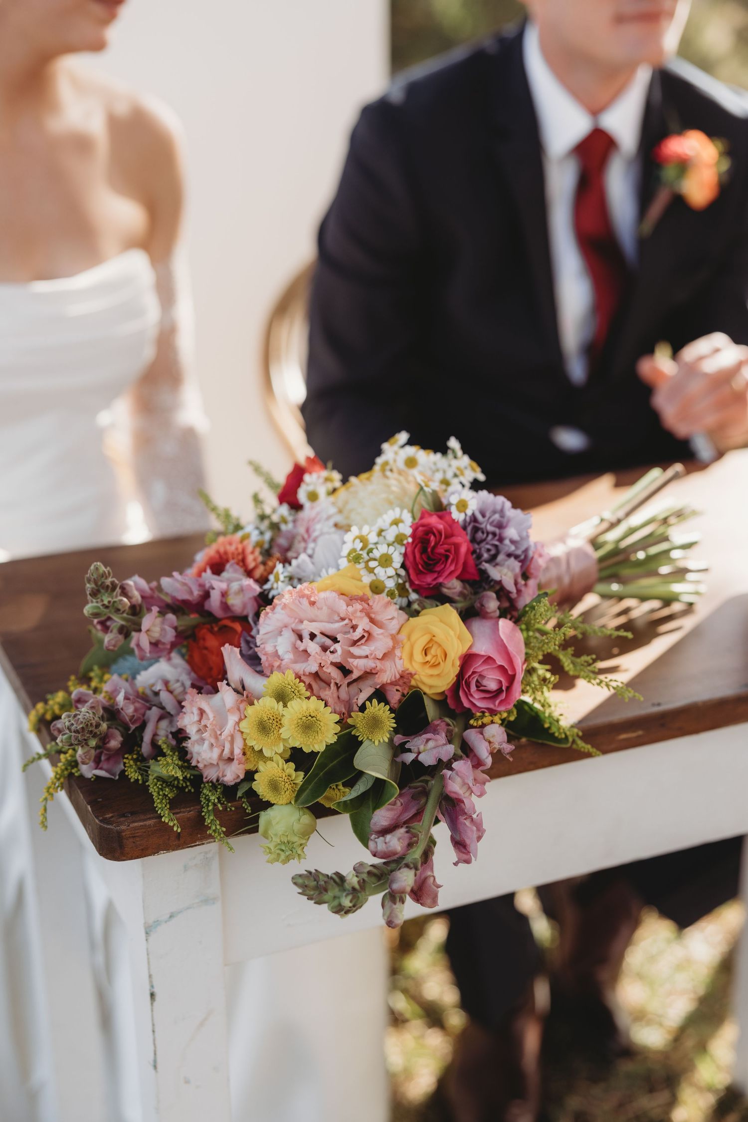 Colorful bridal bouquet with pink, yellow, and red flowers resting on a white wooden chair at a wedding ceremony.