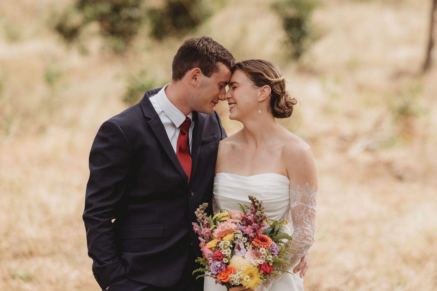 Romantic wedding couple sharing tender moment, bride holding vibrant colorful bouquet in sun-drenched outdoor setting.