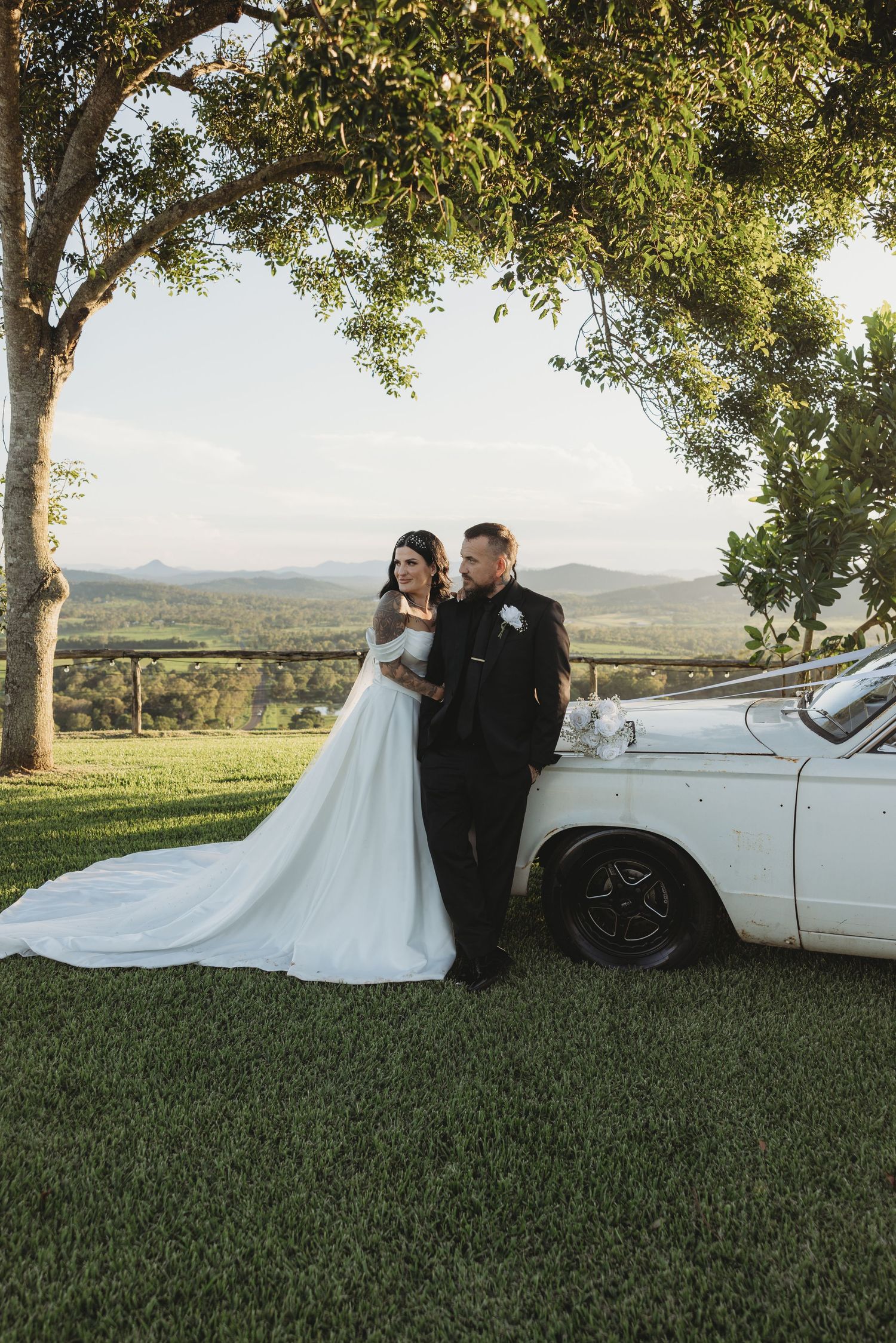 A wedding couple poses next to a vintage white car with mountains and trees in the background.