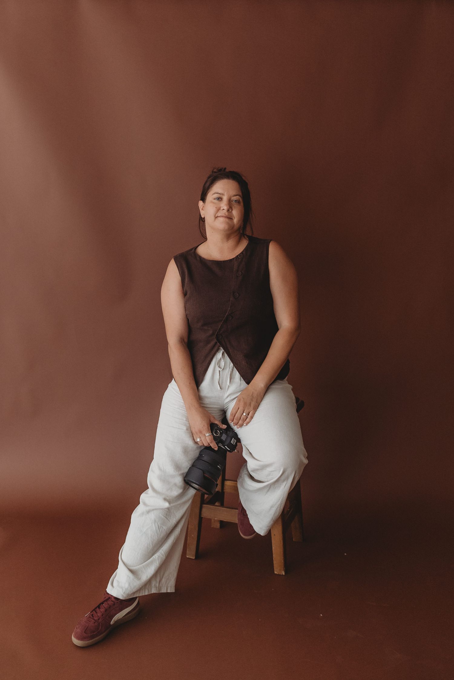 Portrait of photographer sitting on wooden stool with camera in studio setting.