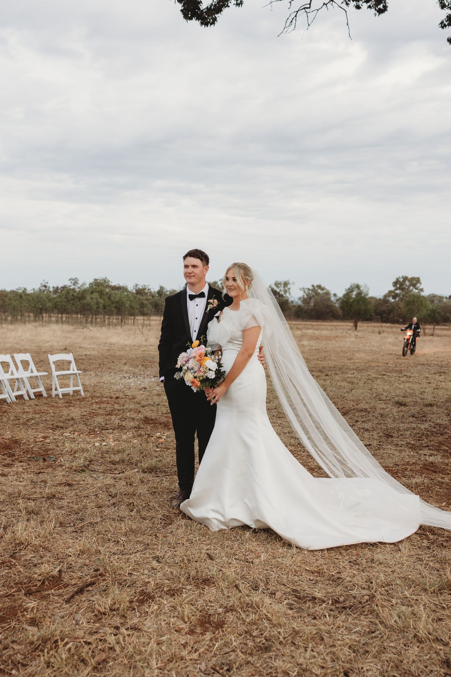 Elegant bride in flowing white dress and groom pose in rustic field with white chairs in background.