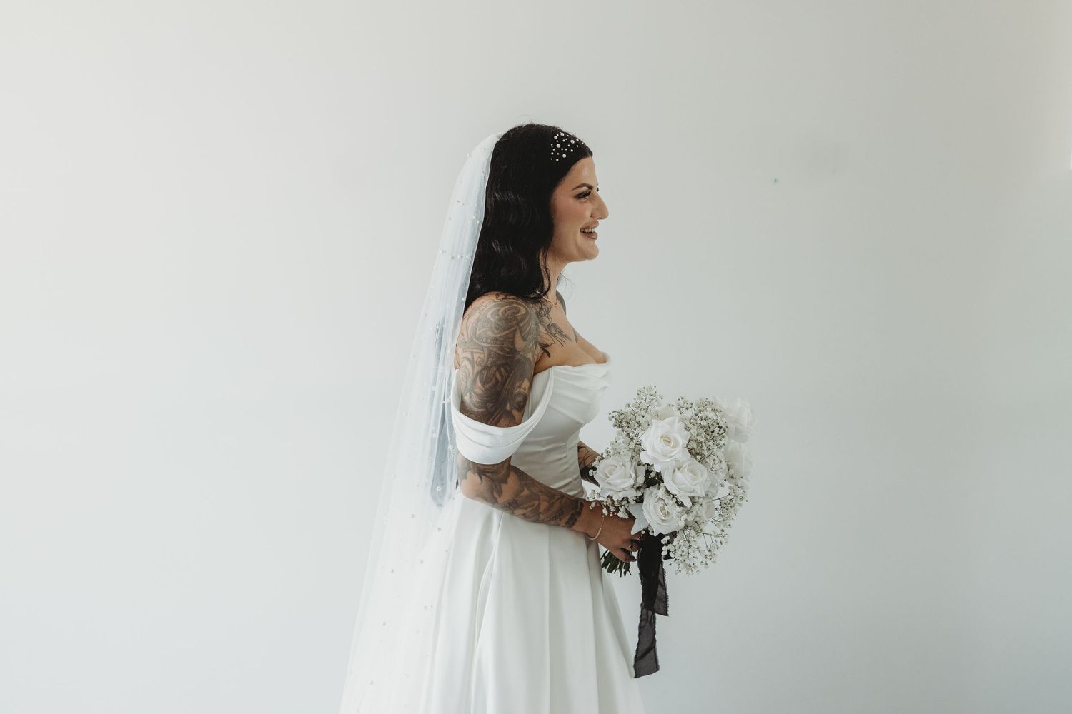 A bride in a white wedding gown holds a bouquet of white flowers against a light gray background.