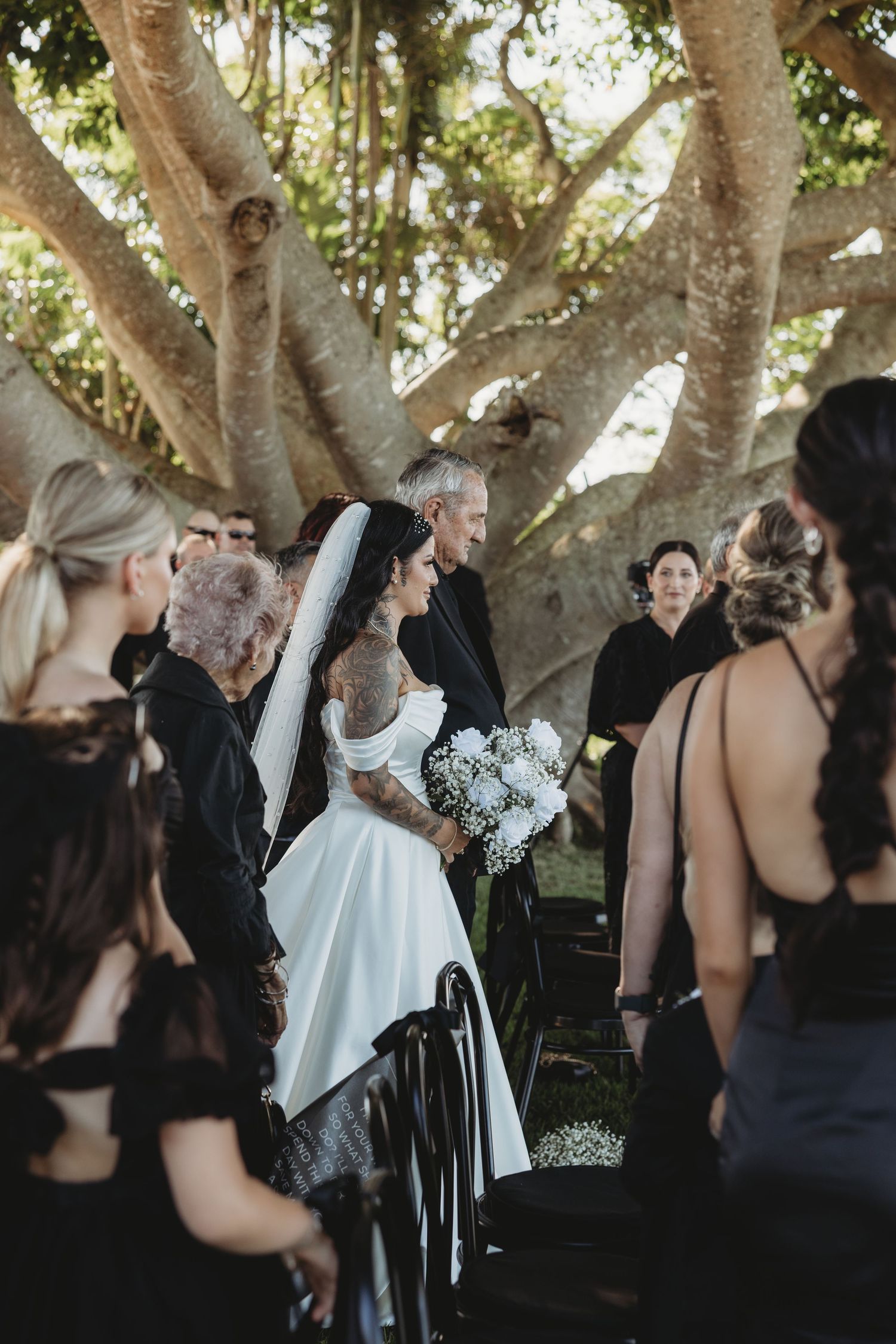 Wedding ceremony under large trees with guests in black dresses and formal attire gathering outdoors.