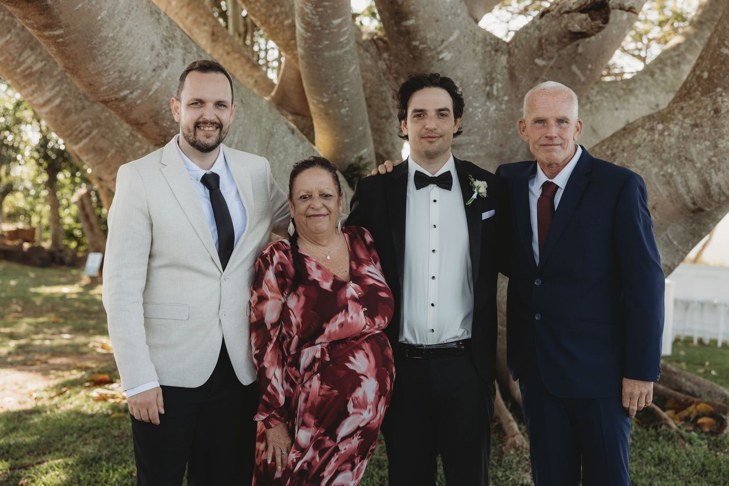 Wedding party members pose for formal photos under tree branches.