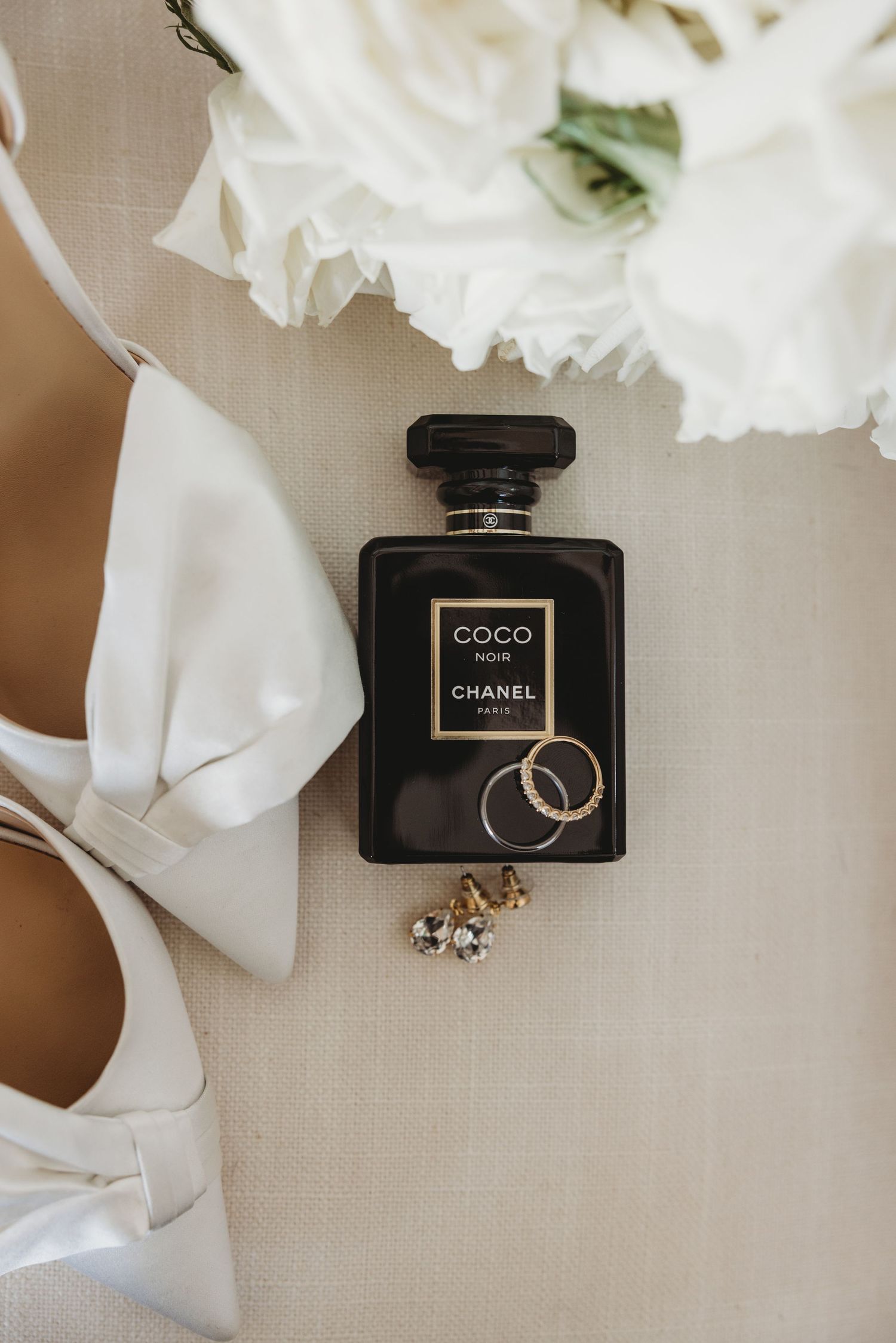 Wedding accessories including Chanel perfume, rings, and white flowers arranged on beige fabric.