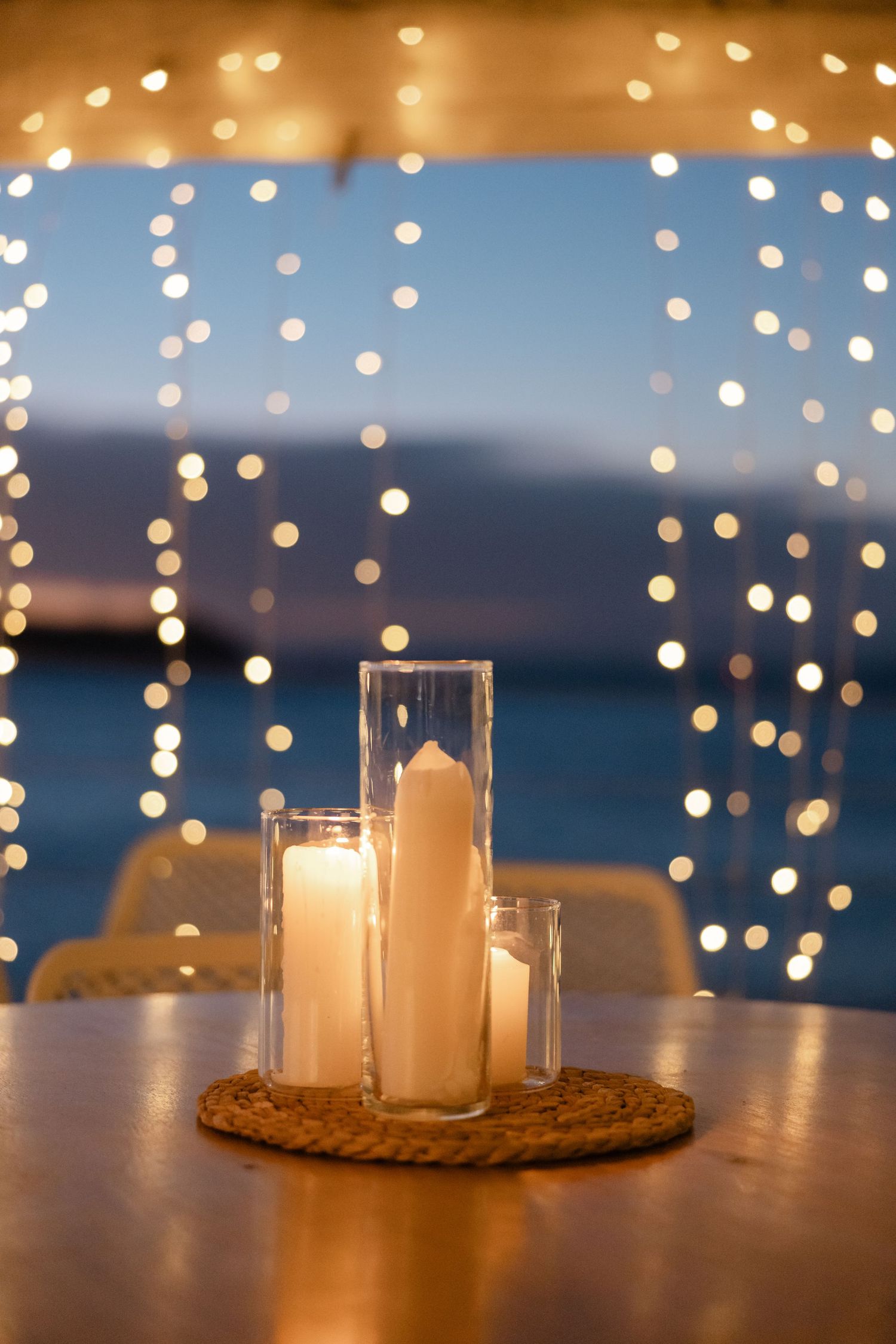 Romantic candlelit table with three pillar candles in glass holders, bokeh string lights, and ocean view at dusk.