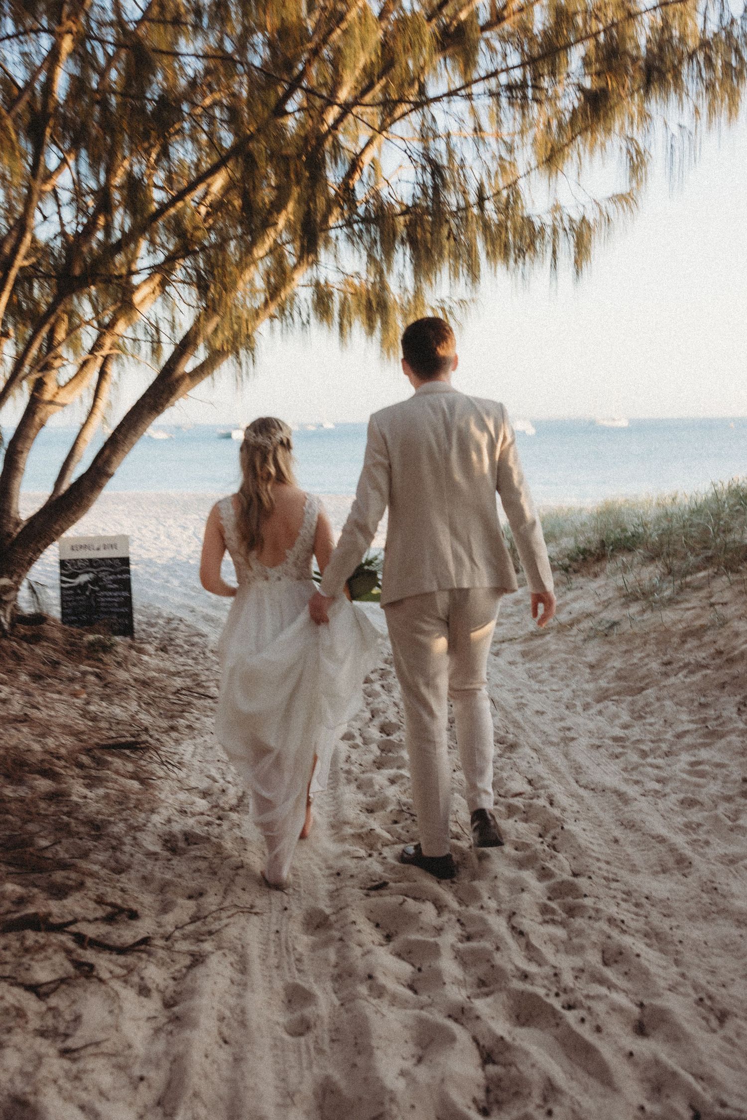 Couple walking along sandy beach path under overhanging trees during sunset.