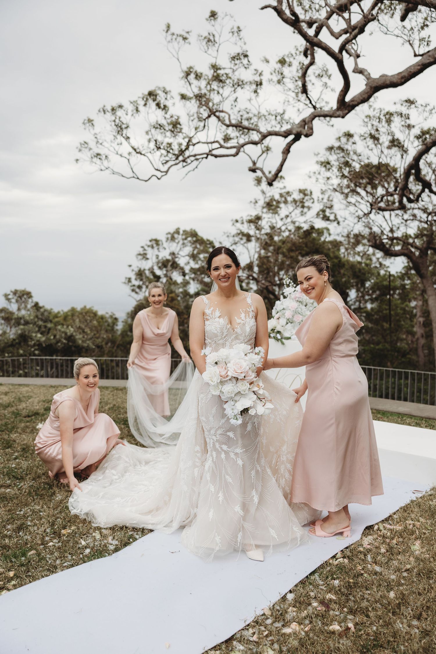 Bridesmaids in blush pink dresses help arrange the bride's lace gown on white fabric outdoors.