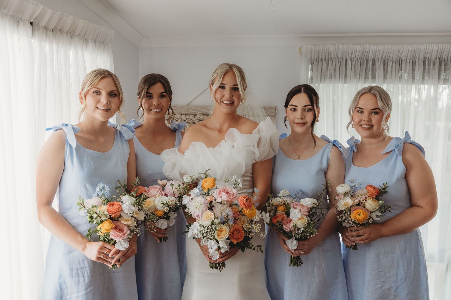 Bridesmaids in powder blue dresses stand together holding peach and orange bouquets.