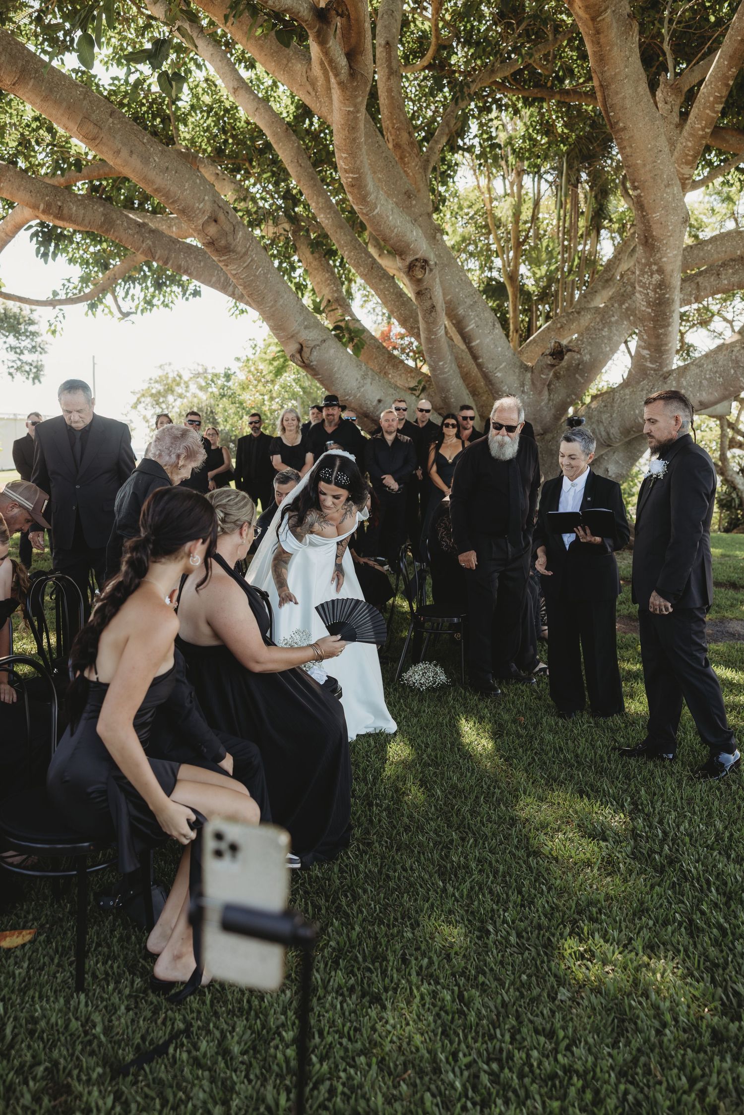 Wedding guests gather under large shady trees during an outdoor ceremony.