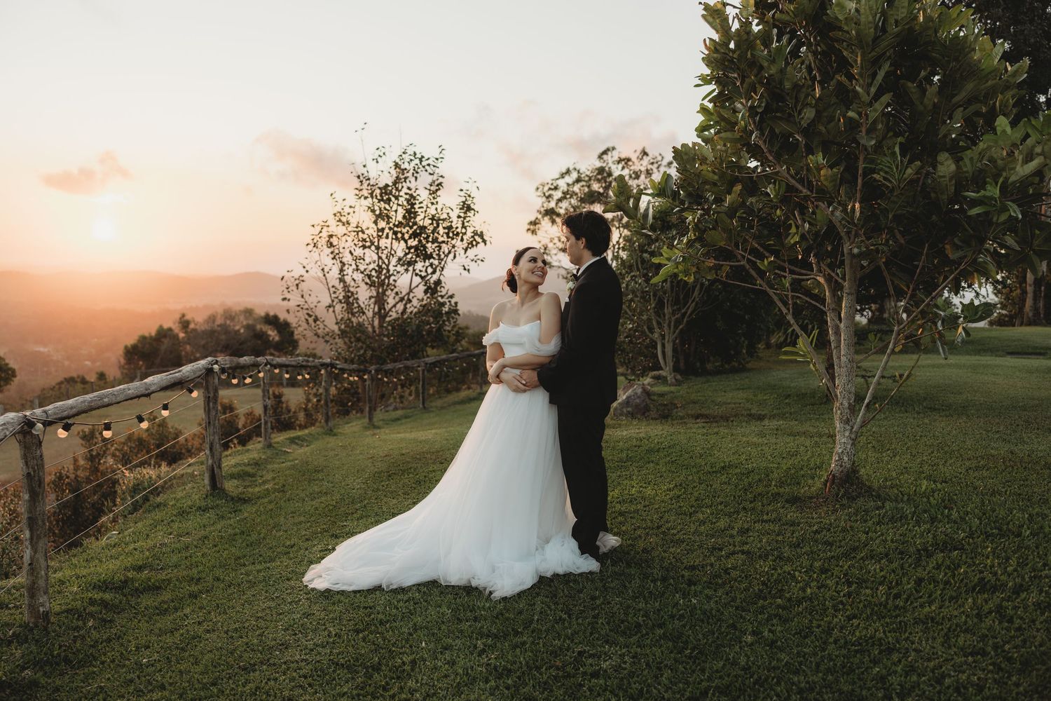 Newlyweds share a romantic moment at sunset on a hilltop with mountains in the background.