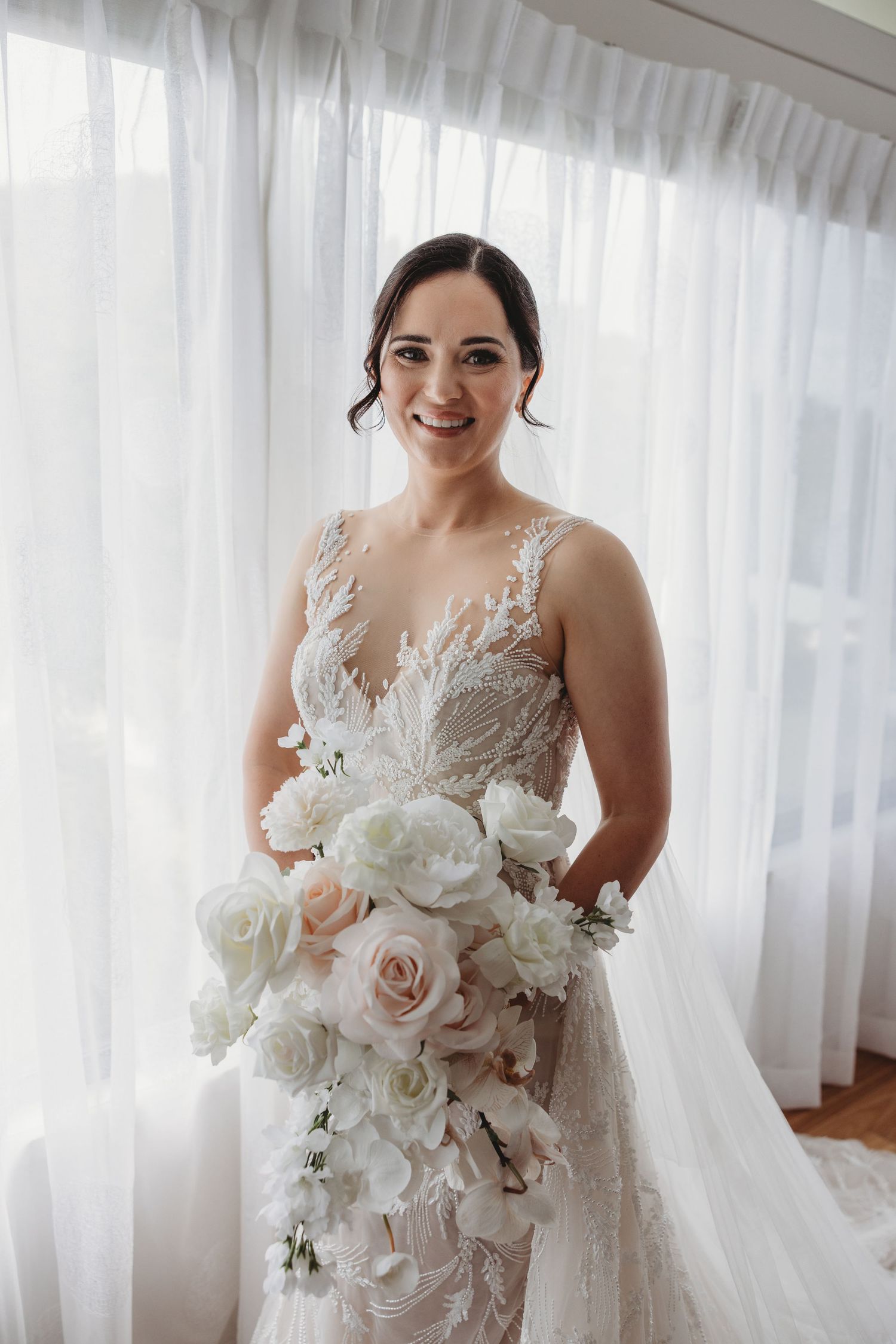 Elegant white bridal bouquet with roses and delicate flowers held against a light curtained background.