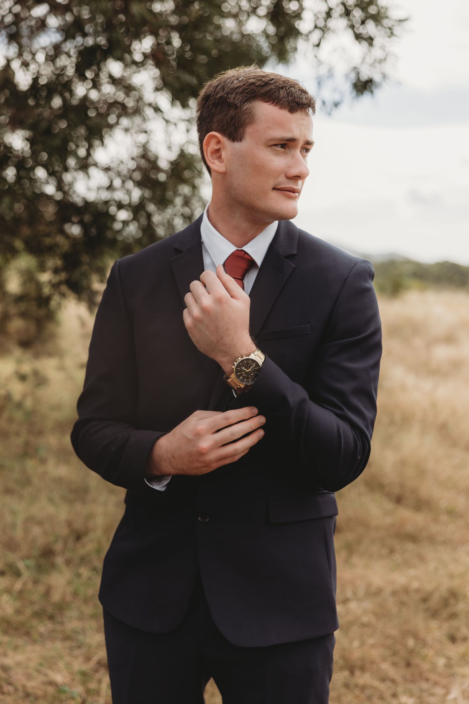 Groom in navy suit and red tie adjusting cufflinks outdoors in a field with trees in background.