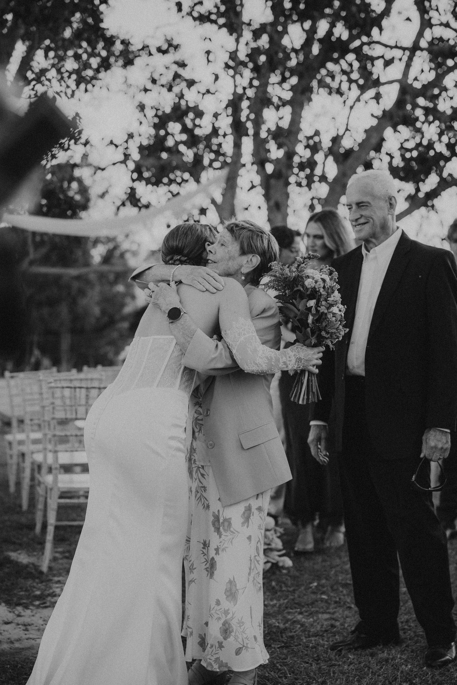 Black and white photo of bride embracing guest at outdoor wedding ceremony while smiling elderly man watches nearby.