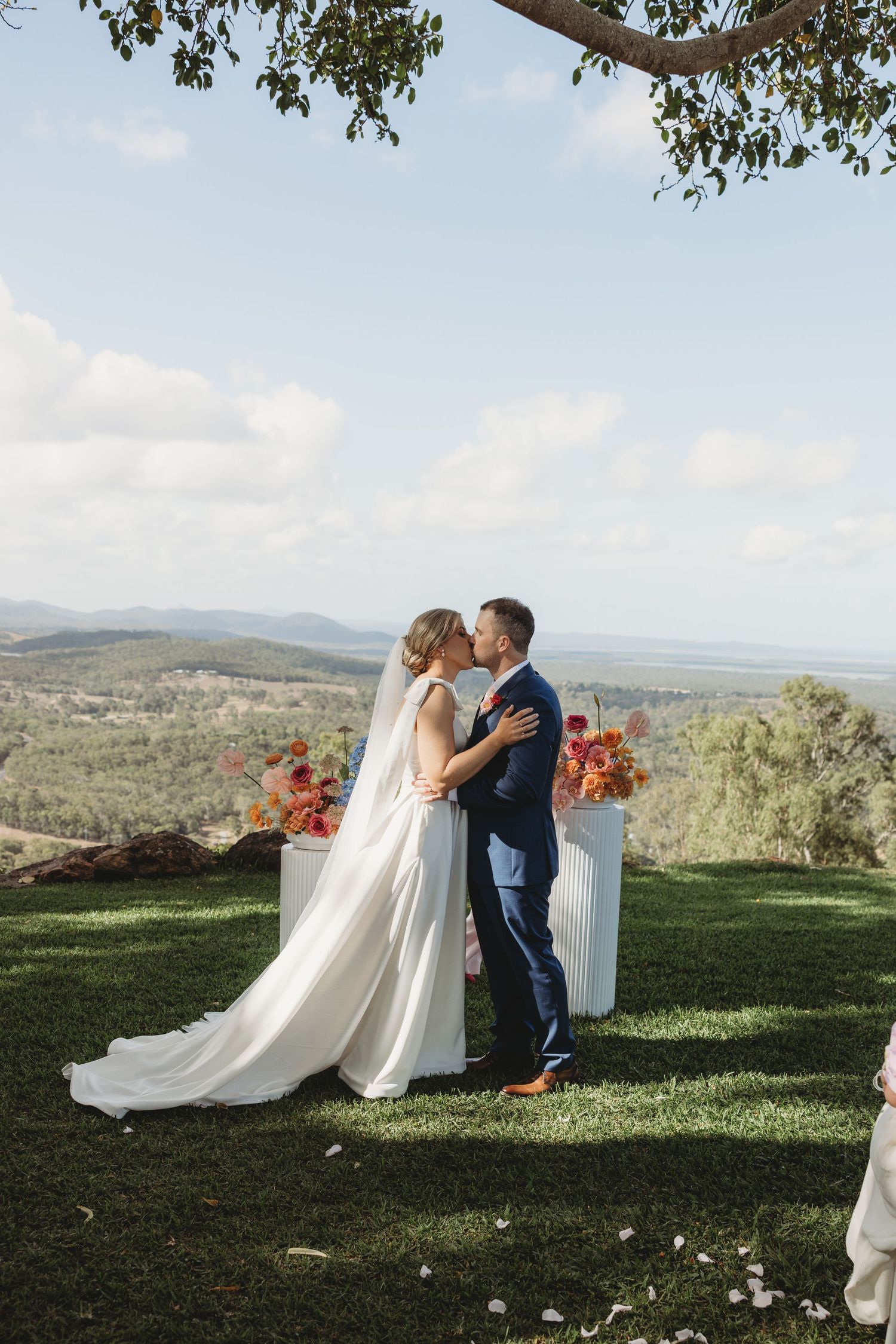 Wedding couple embraces on hilltop overlooking scenic mountain landscape with flowing white dress.
