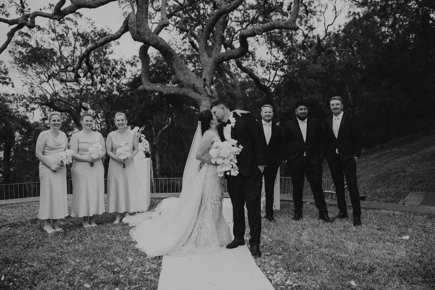 Wedding party poses outdoors in formal attire with bridesmaids in white dresses and groomsmen in dark suits.