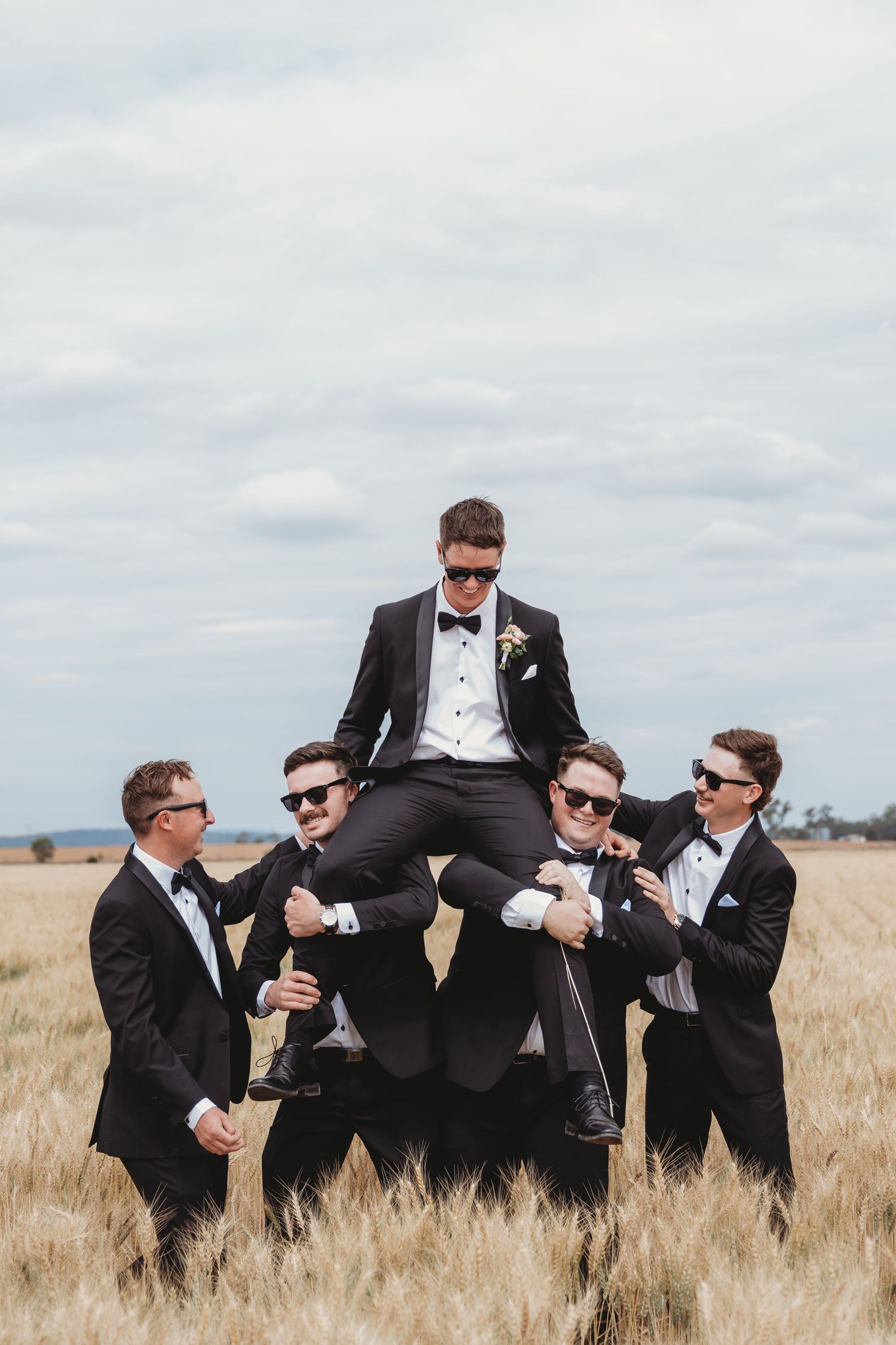 Wedding party lifting groomsman in wheat field with dramatic cloudy sky.