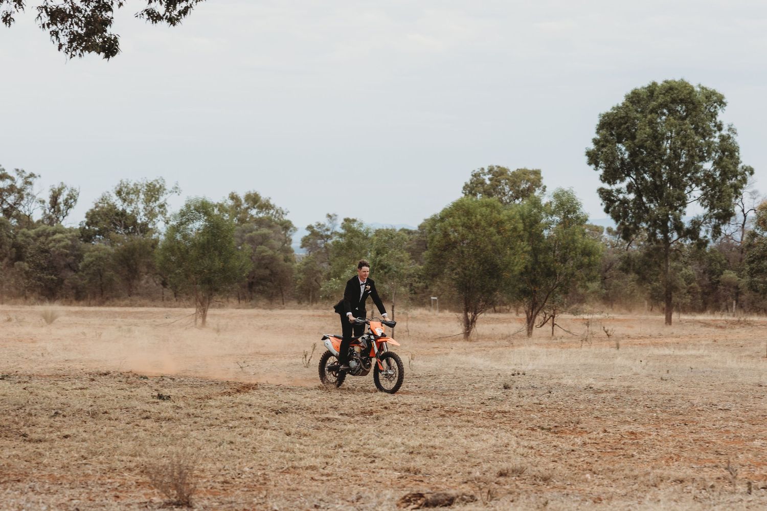 A dirt bike rider traverses across a dusty outback landscape with scattered trees in the background.