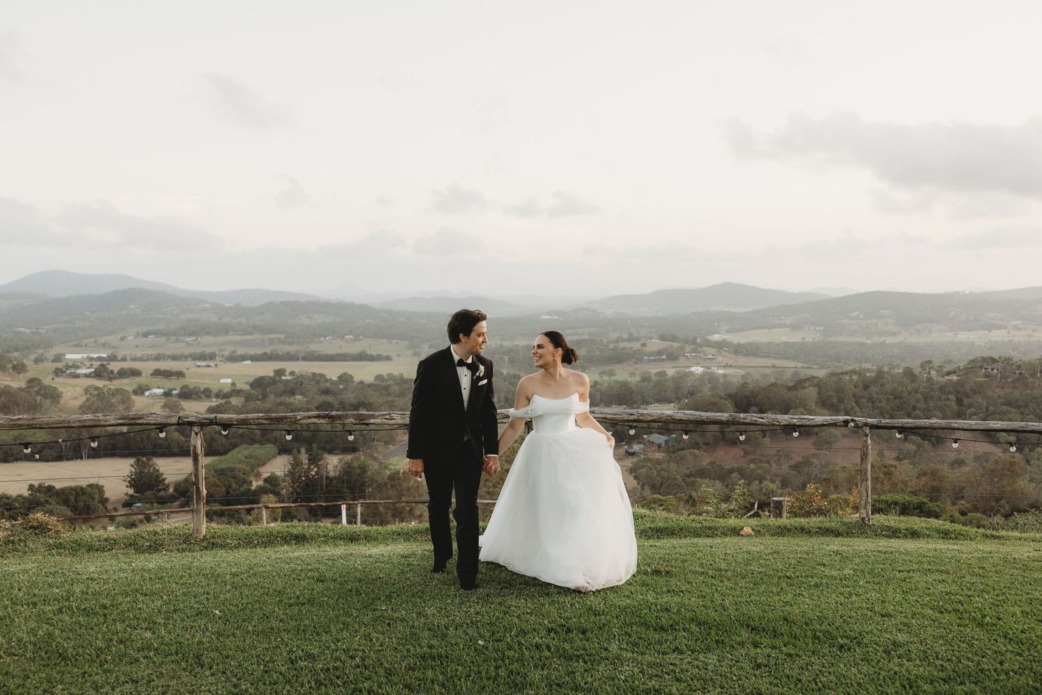 Wedding couple stands together on a grassy hilltop with a panoramic valley view behind them.
