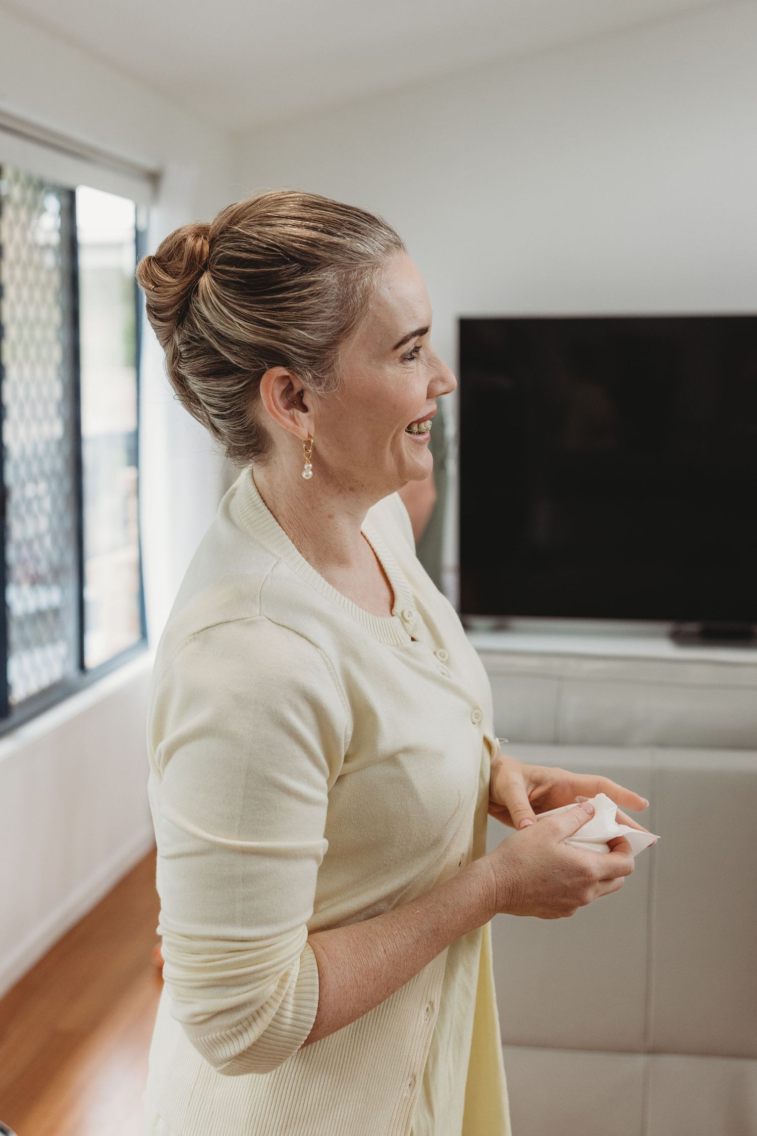 A person in a cream-colored top smiles and gestures while standing in a bright interior room.