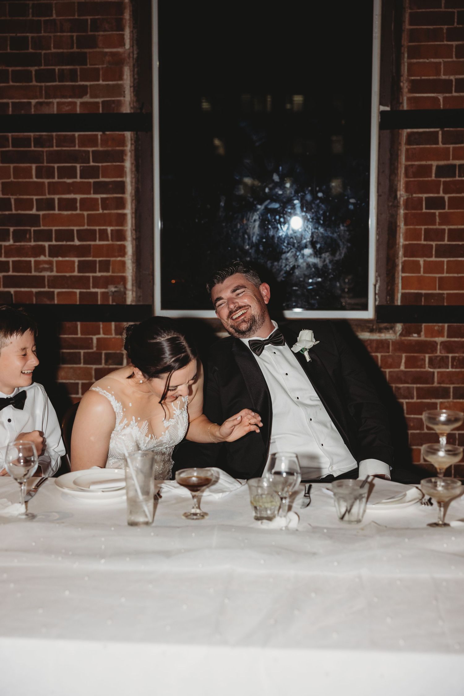 Wedding party seated at long table against brick wall during reception celebration.