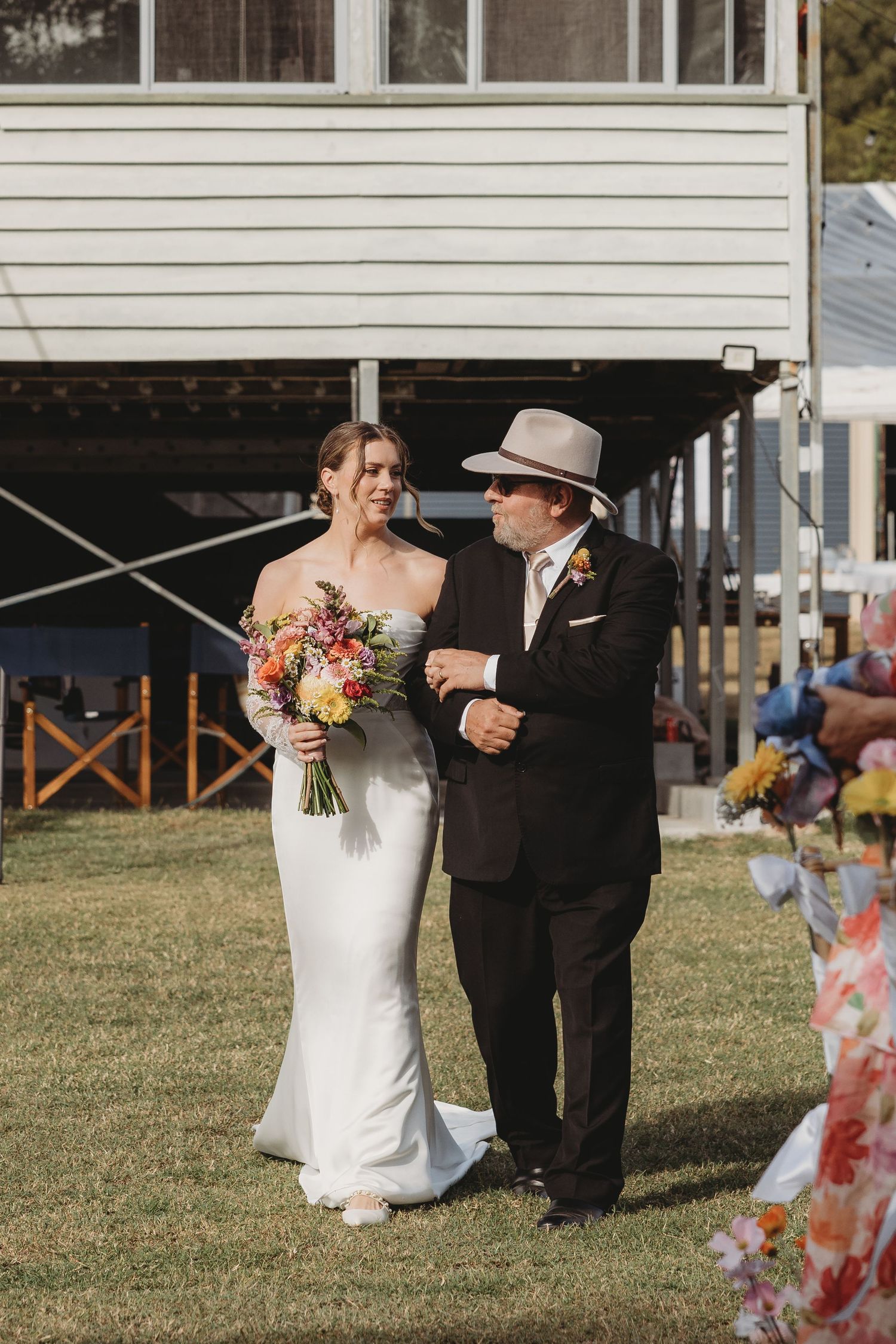 Bride walks down the aisle with escort holding colorful bouquet at outdoor wedding ceremony.