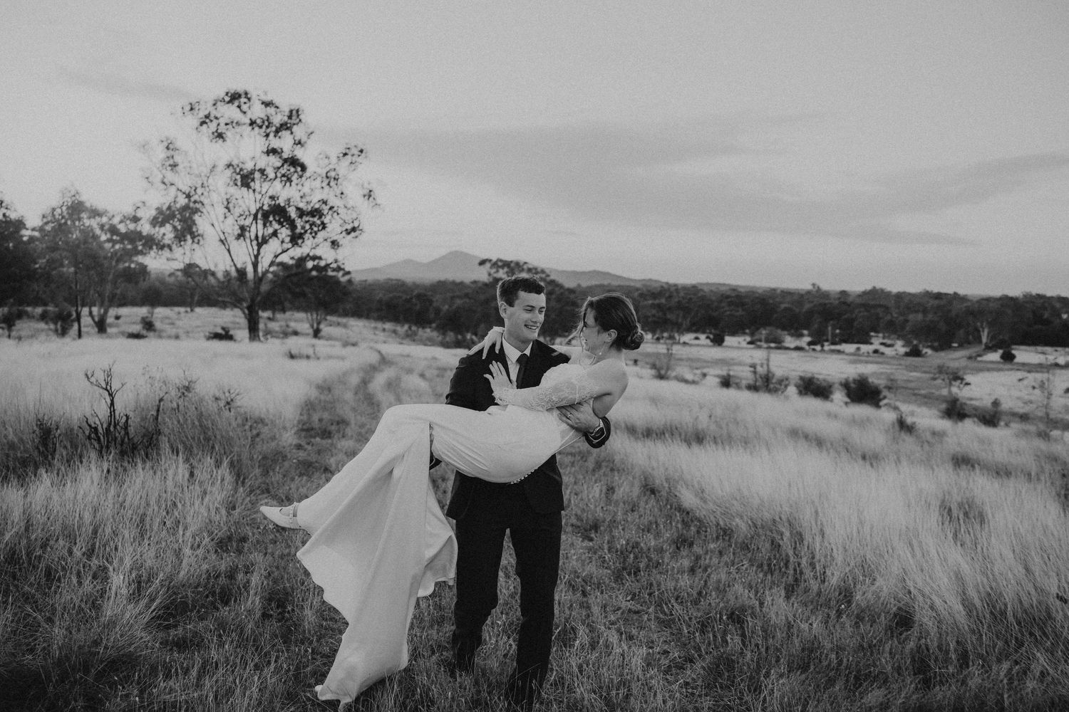 Groom carrying bride in a field at dusk, black and white wedding photo with mountains in background.