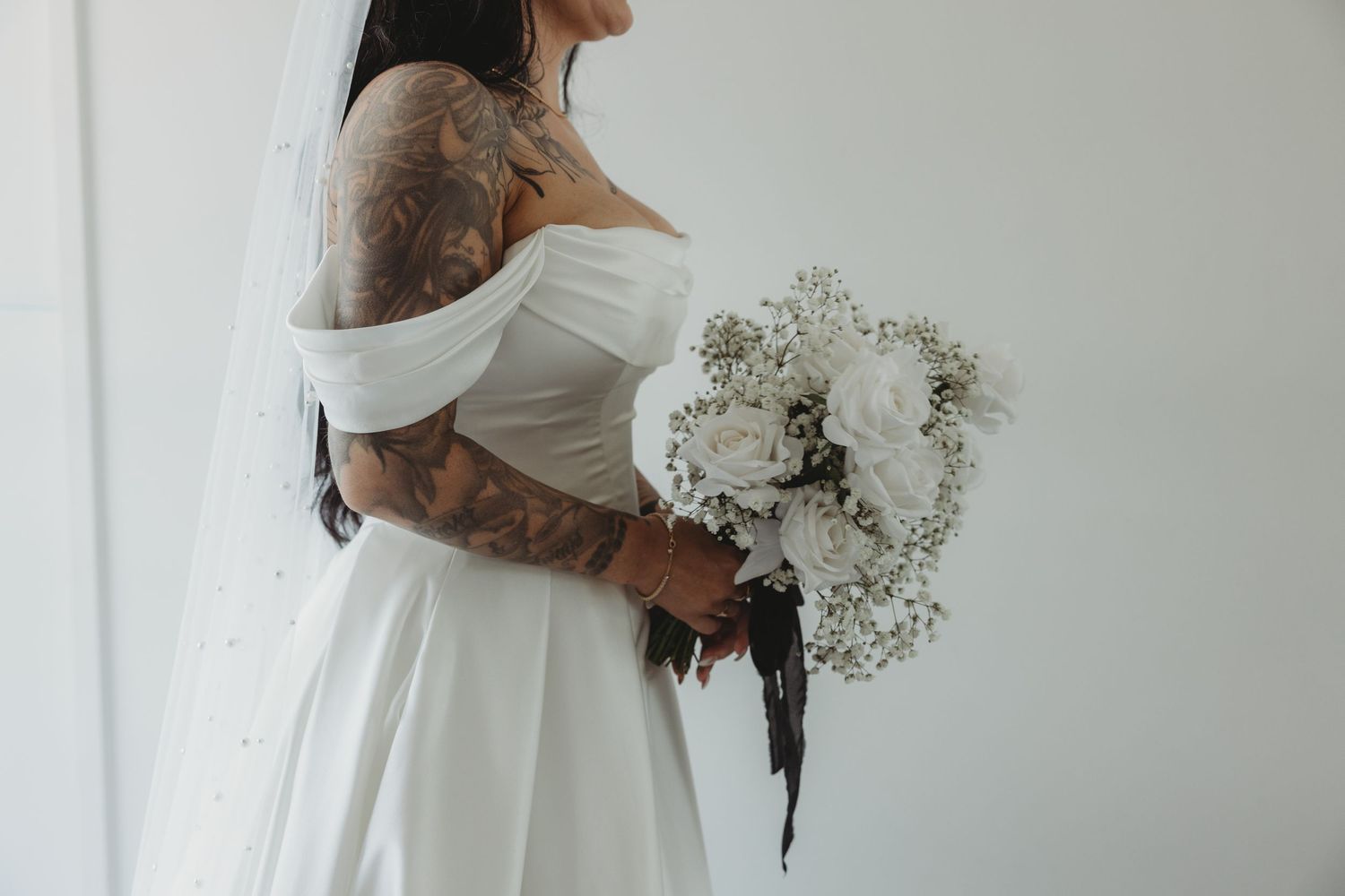 A bride holding a white baby's breath bouquet stands in an elegant white wedding dress against a light background.