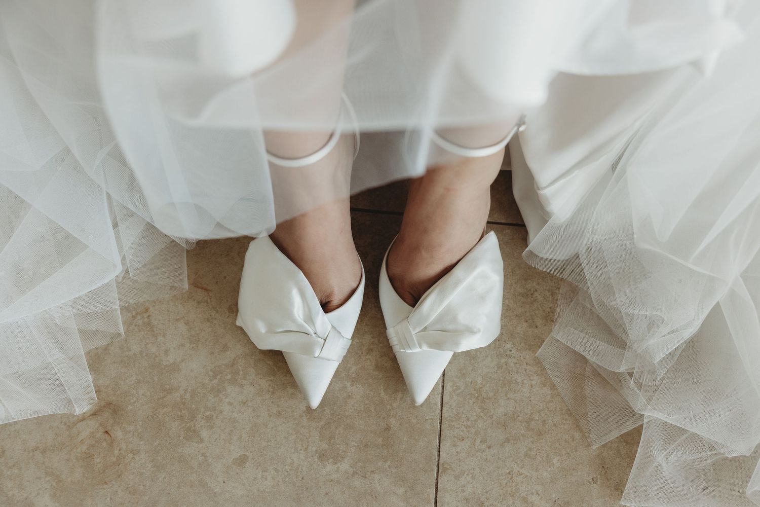 Close-up of white bridal shoes and wedding dress hem on light flooring.