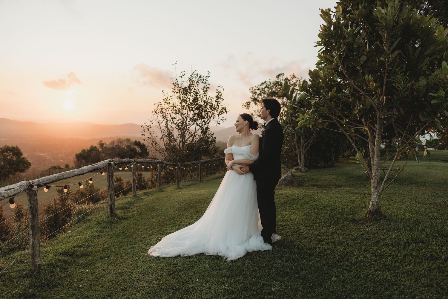 A sunset wedding portrait featuring newlyweds embracing on a grassy hillside with rustic fencing.