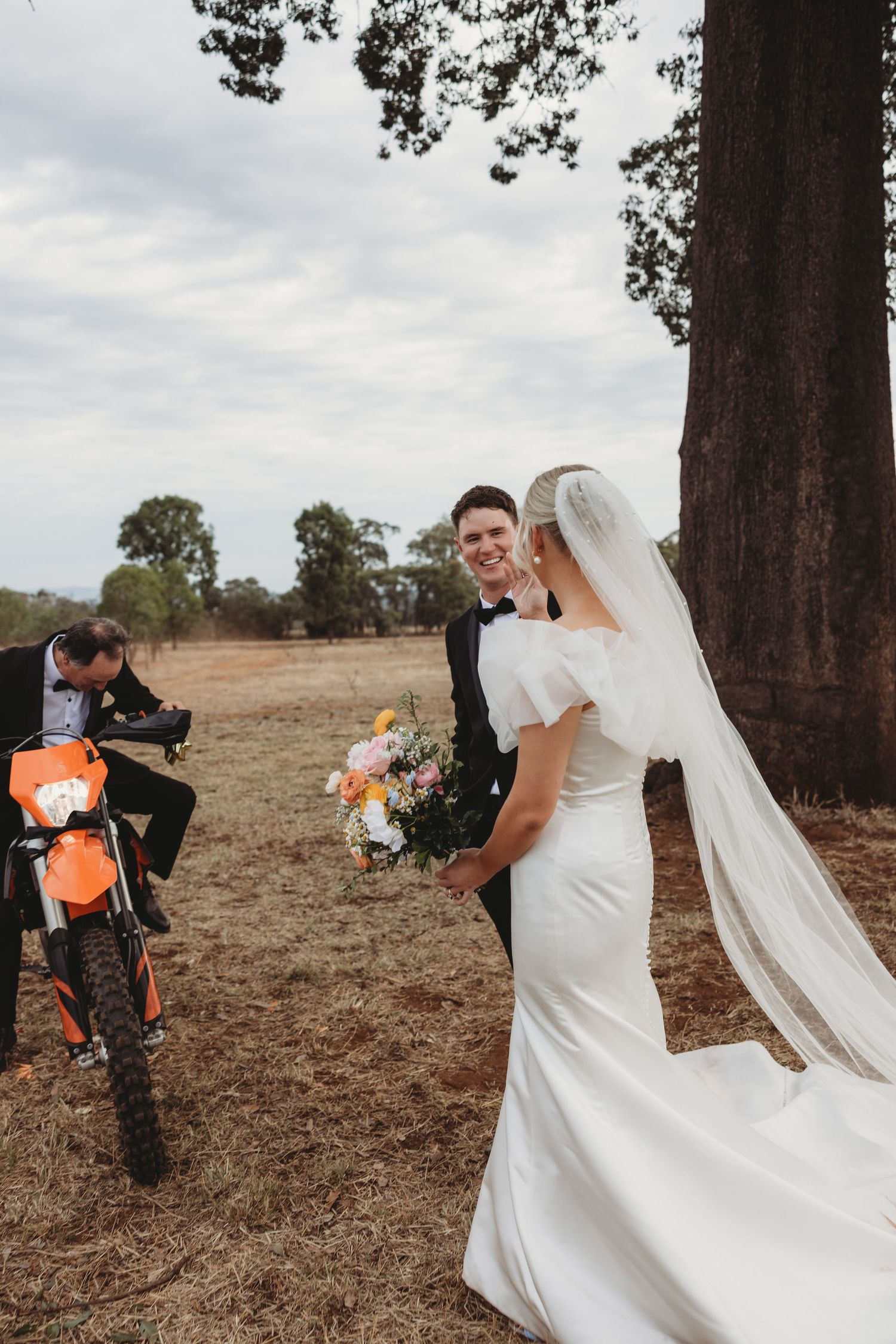 Wedding couple poses with orange dirt bike in rustic outdoor setting.