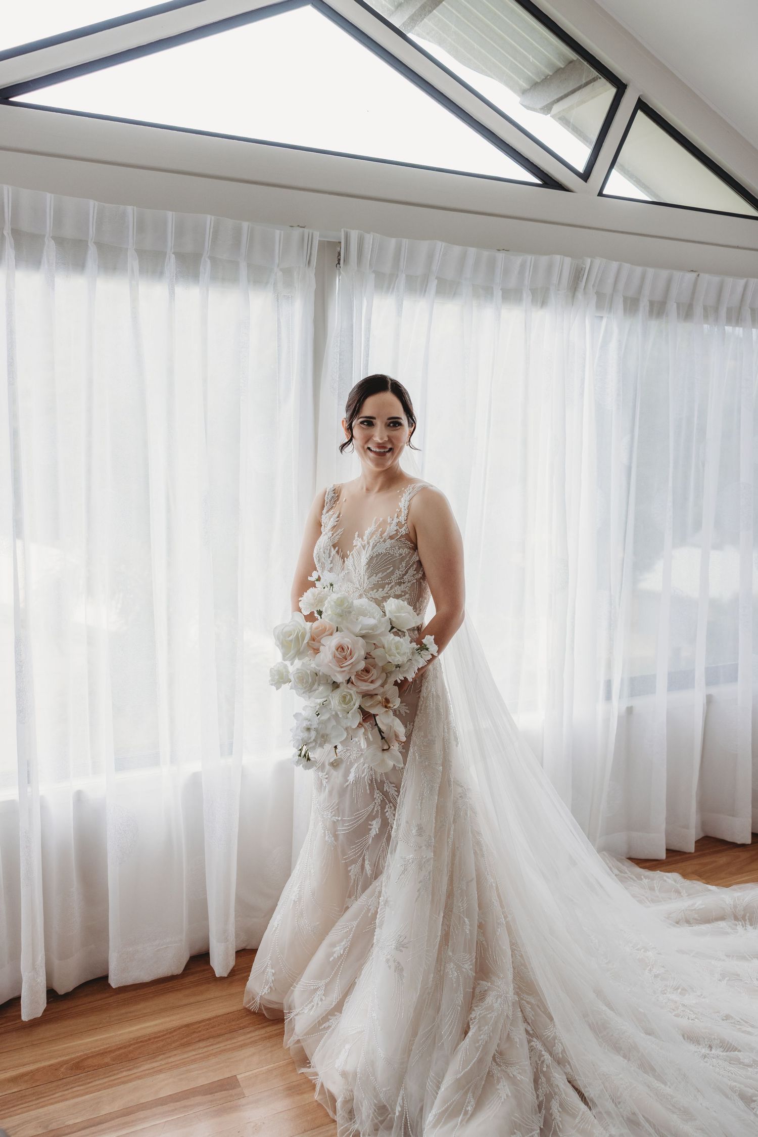 A bride stands in a luxurious white lace wedding gown with flowing train against sheer curtains in a sunlit room.