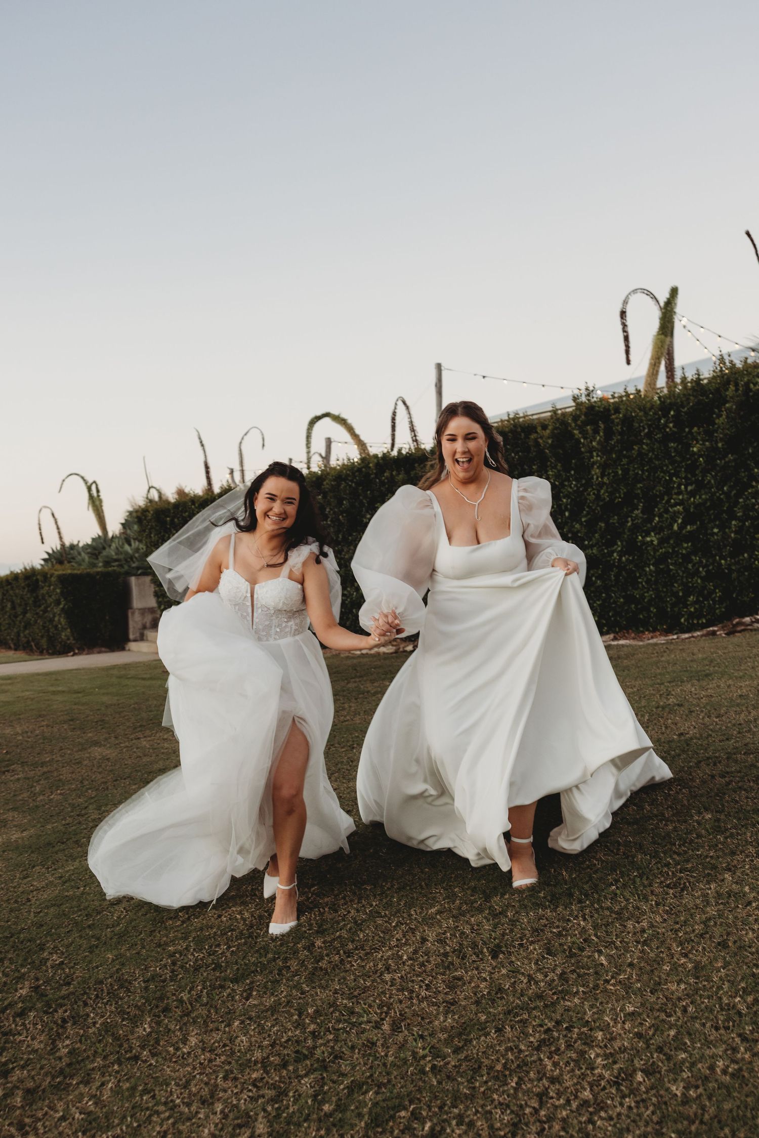 Two people in white dresses playfully run through a grassy field at sunset.