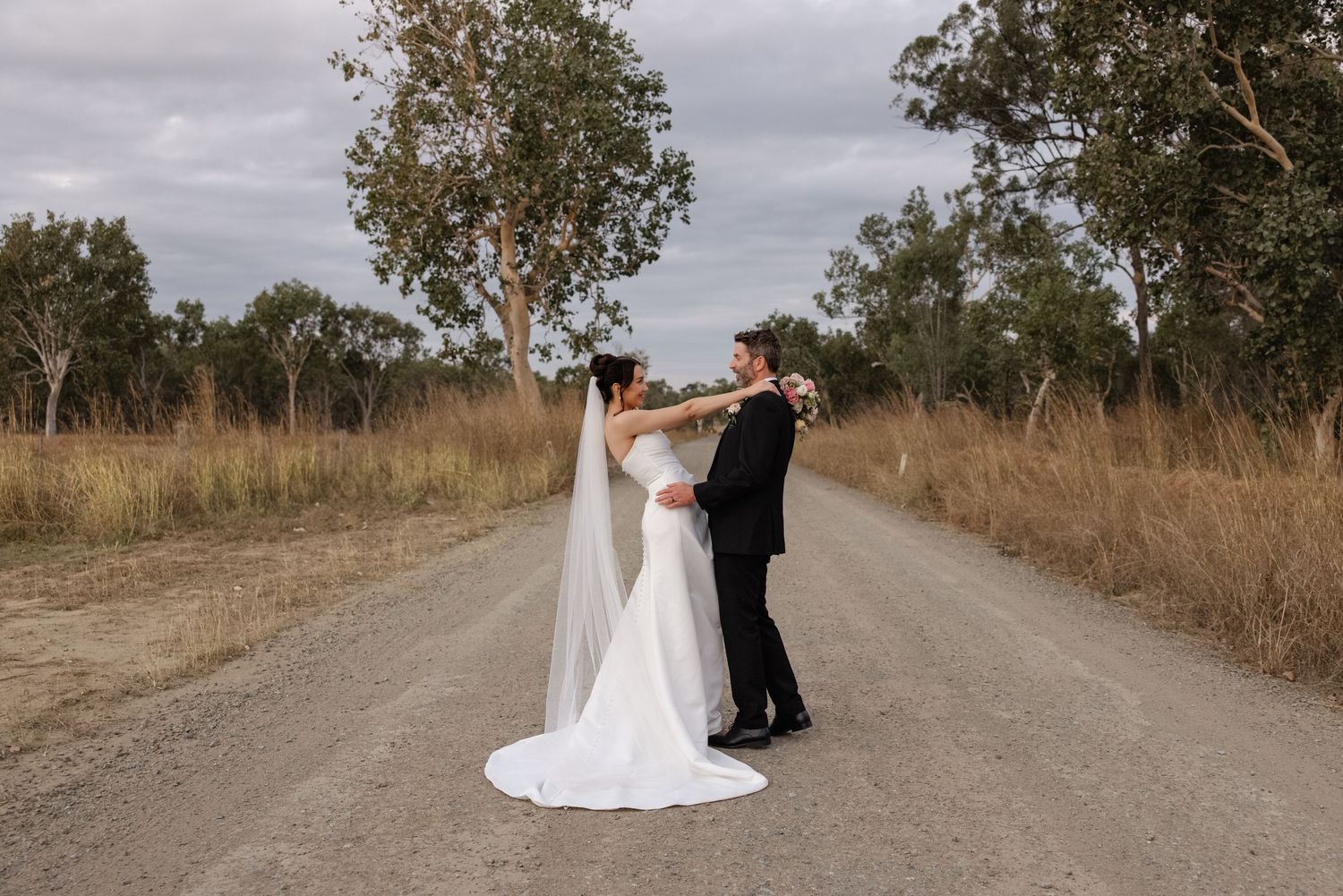 Bride and groom dancing on a rural gravel road surrounded by golden grass and eucalyptus trees on their wedding day.
